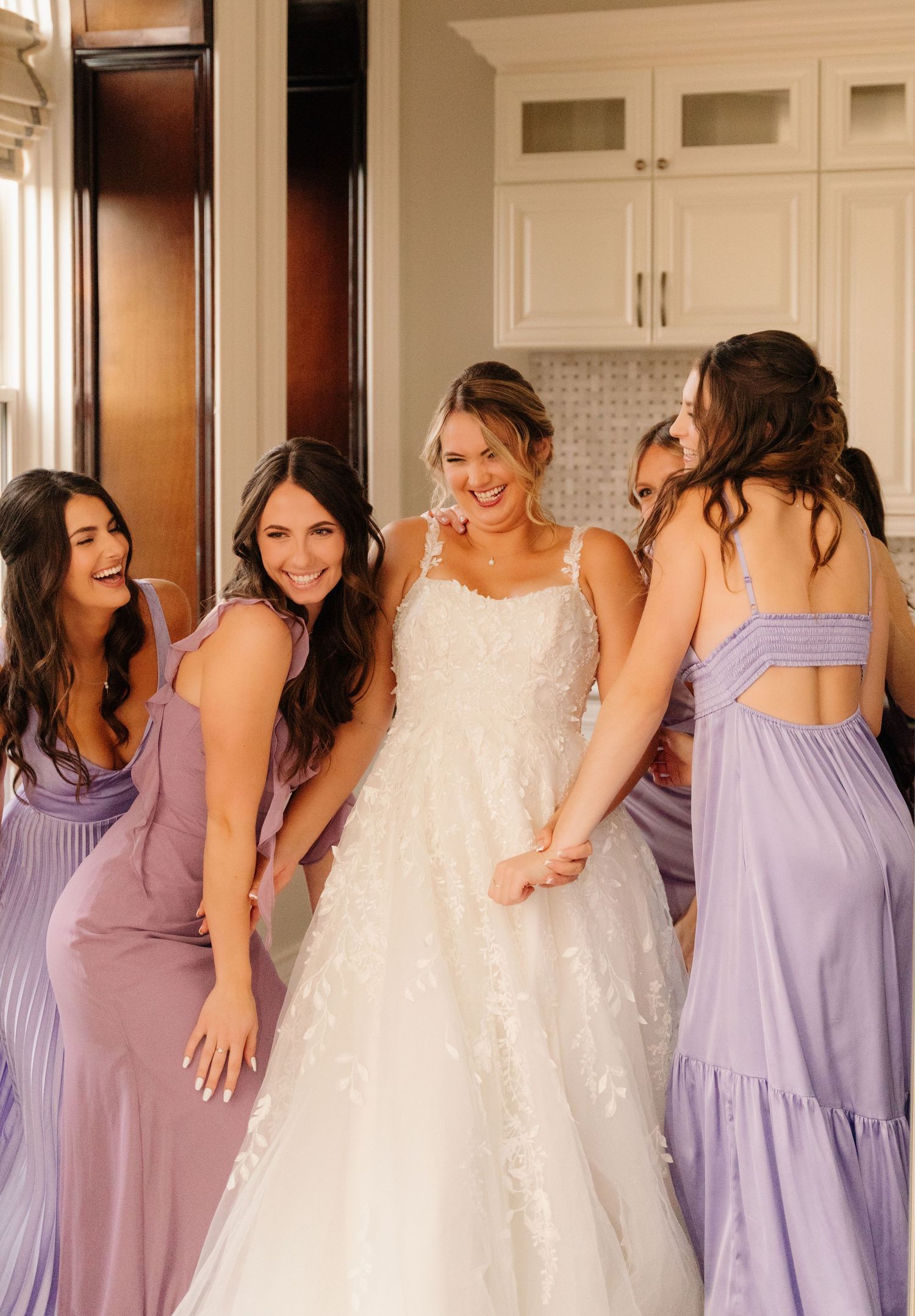 Bridal group in purple and lavender dresses posing together in a modern kitchen.