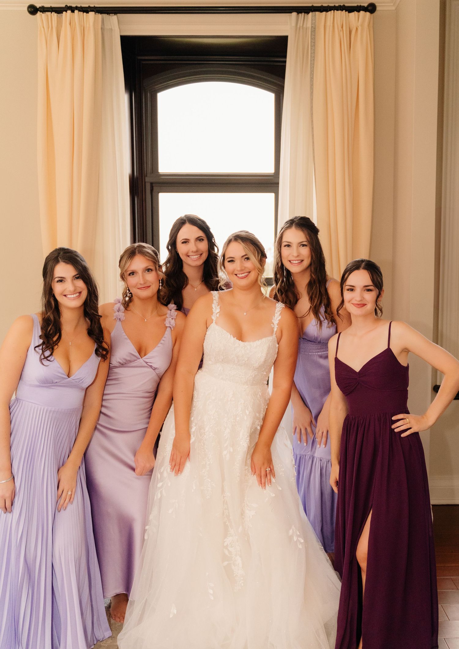 Formal bridal party portrait featuring lavender and burgundy dresses against cream curtains.