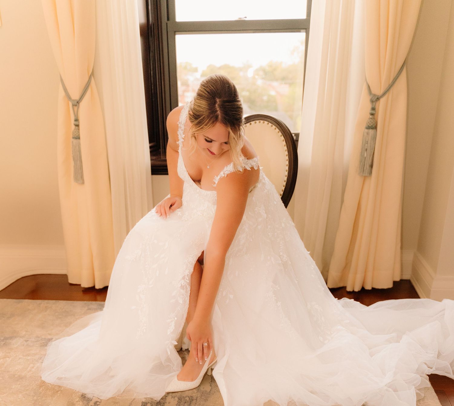 A wedding gown with intricate lace detailing flows elegantly around a seated figure by a bright window.