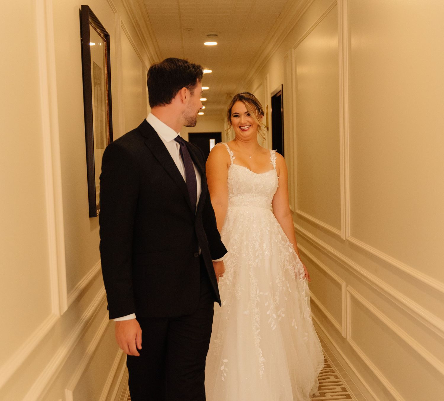 A bride in a white lace dress and groom in black suit walk down an elegant hotel hallway.