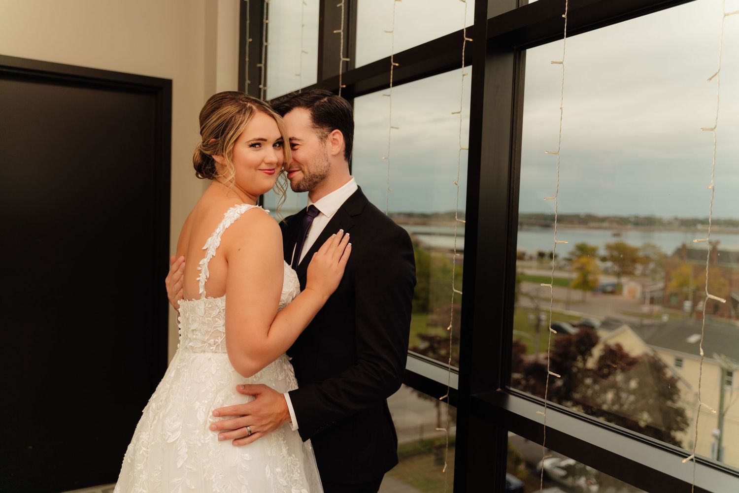 A romantic wedding couple embraces by large windows overlooking a coastal view.