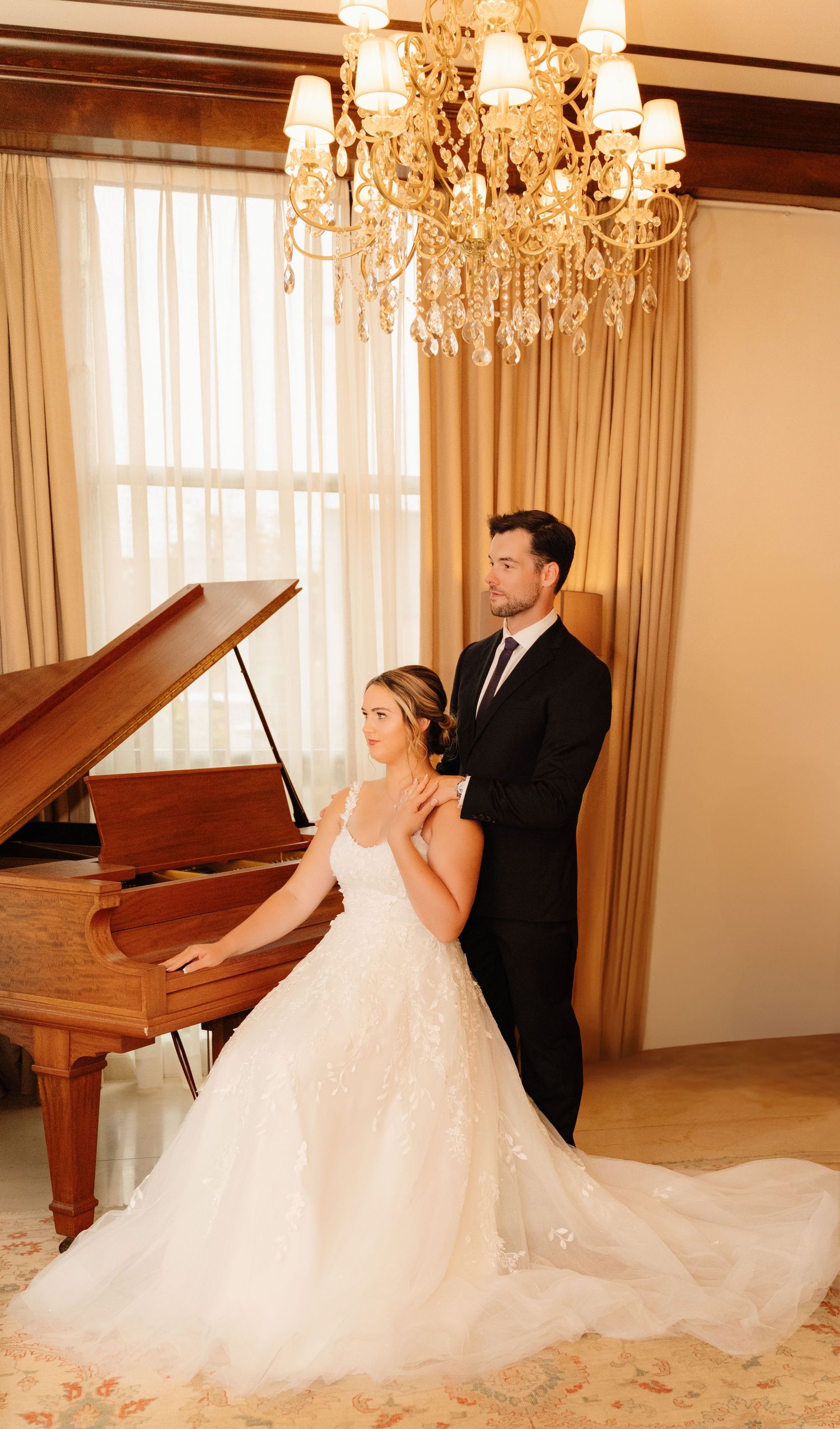 Elegant wedding couple poses by grand piano under crystal chandelier in formal room.