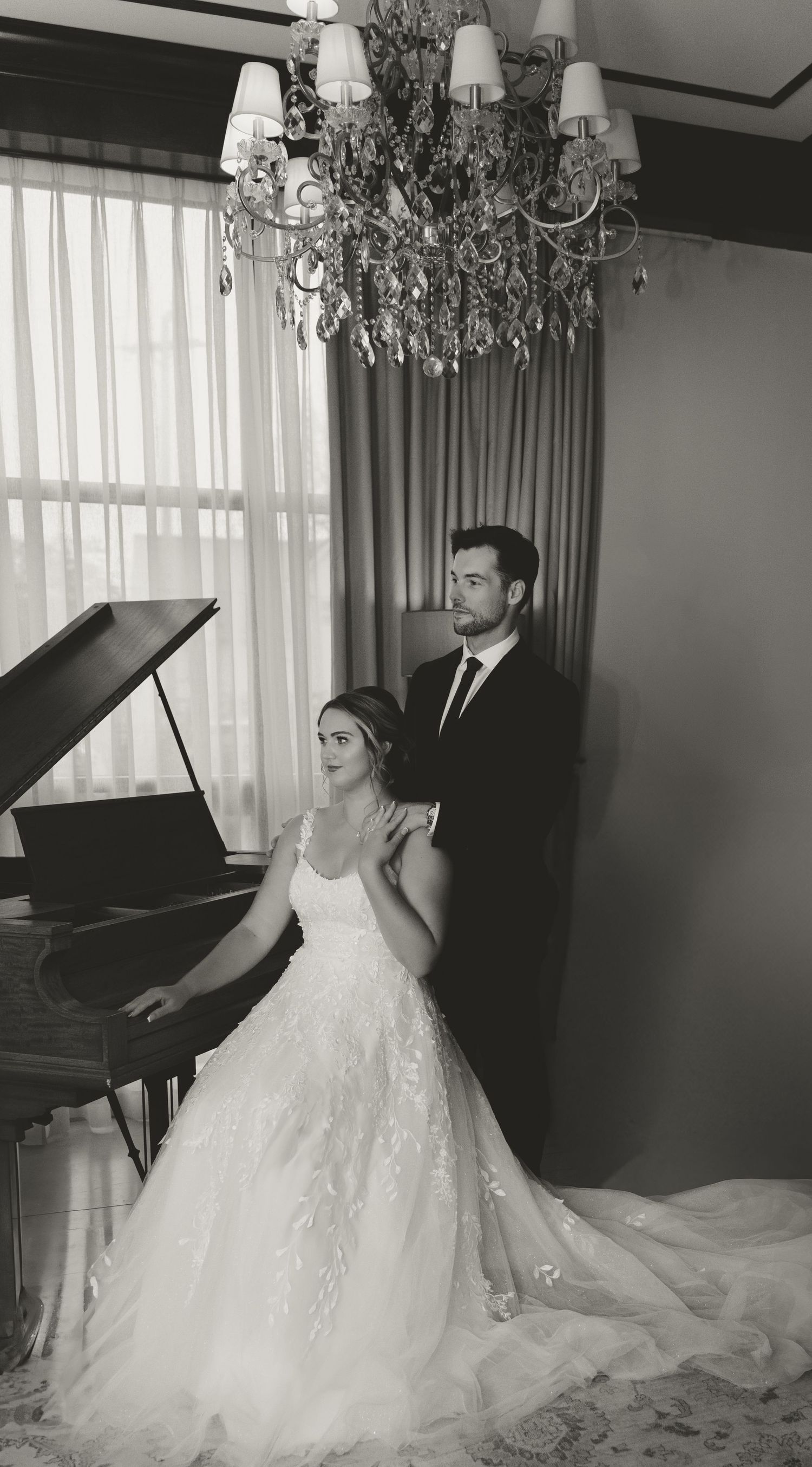 A black and white wedding photo showing a couple posing by a grand piano under an elegant crystal chandelier.