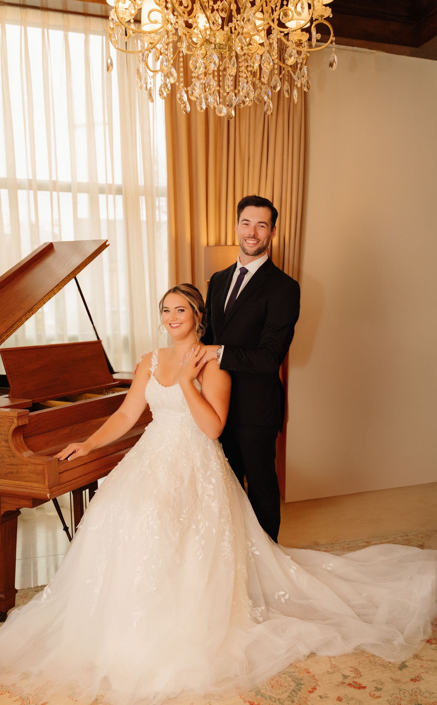 A wedding couple poses together by a piano in an elegant room with a crystal chandelier.