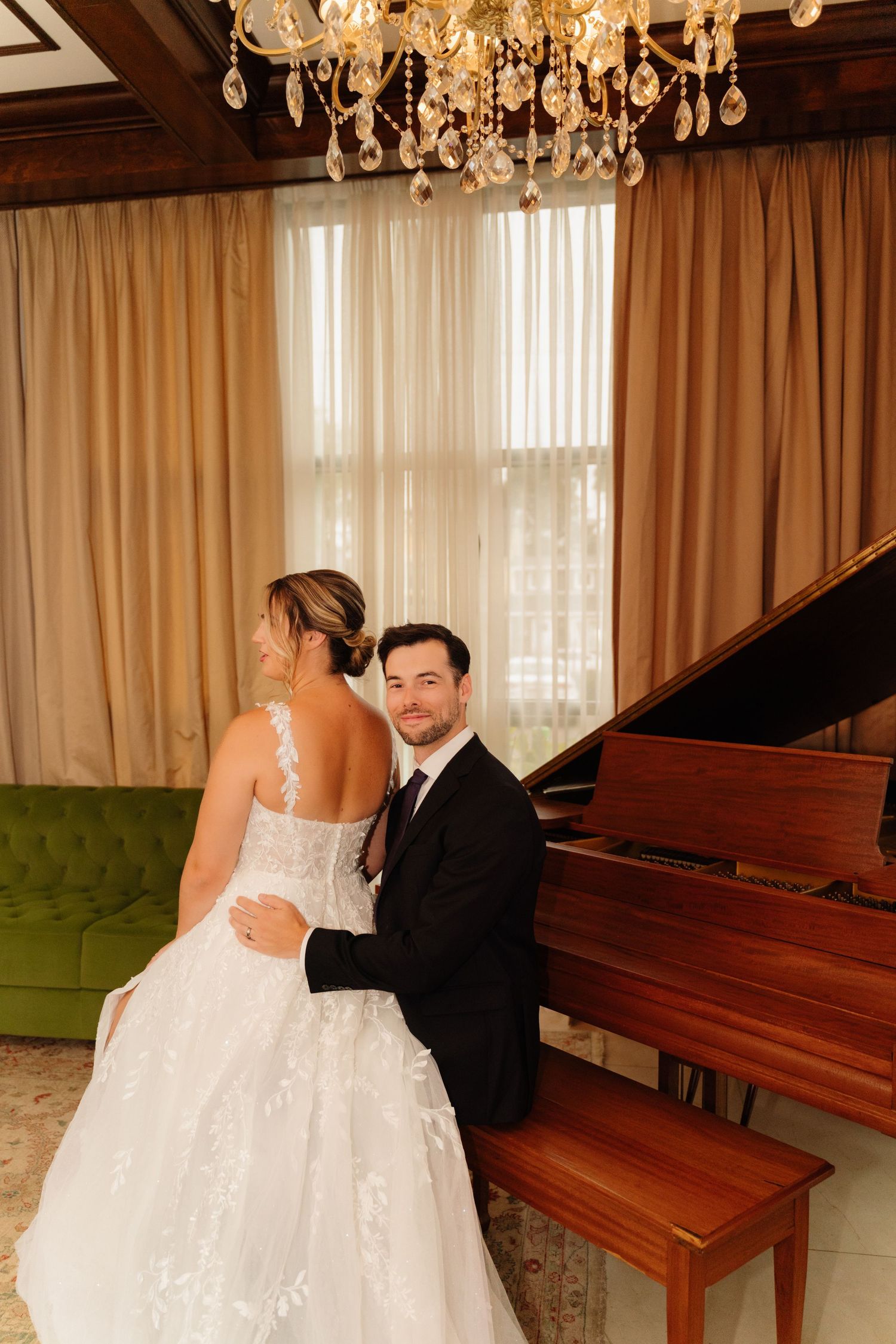 Newlywed couple shares an intimate moment at a piano bench under an ornate chandelier.