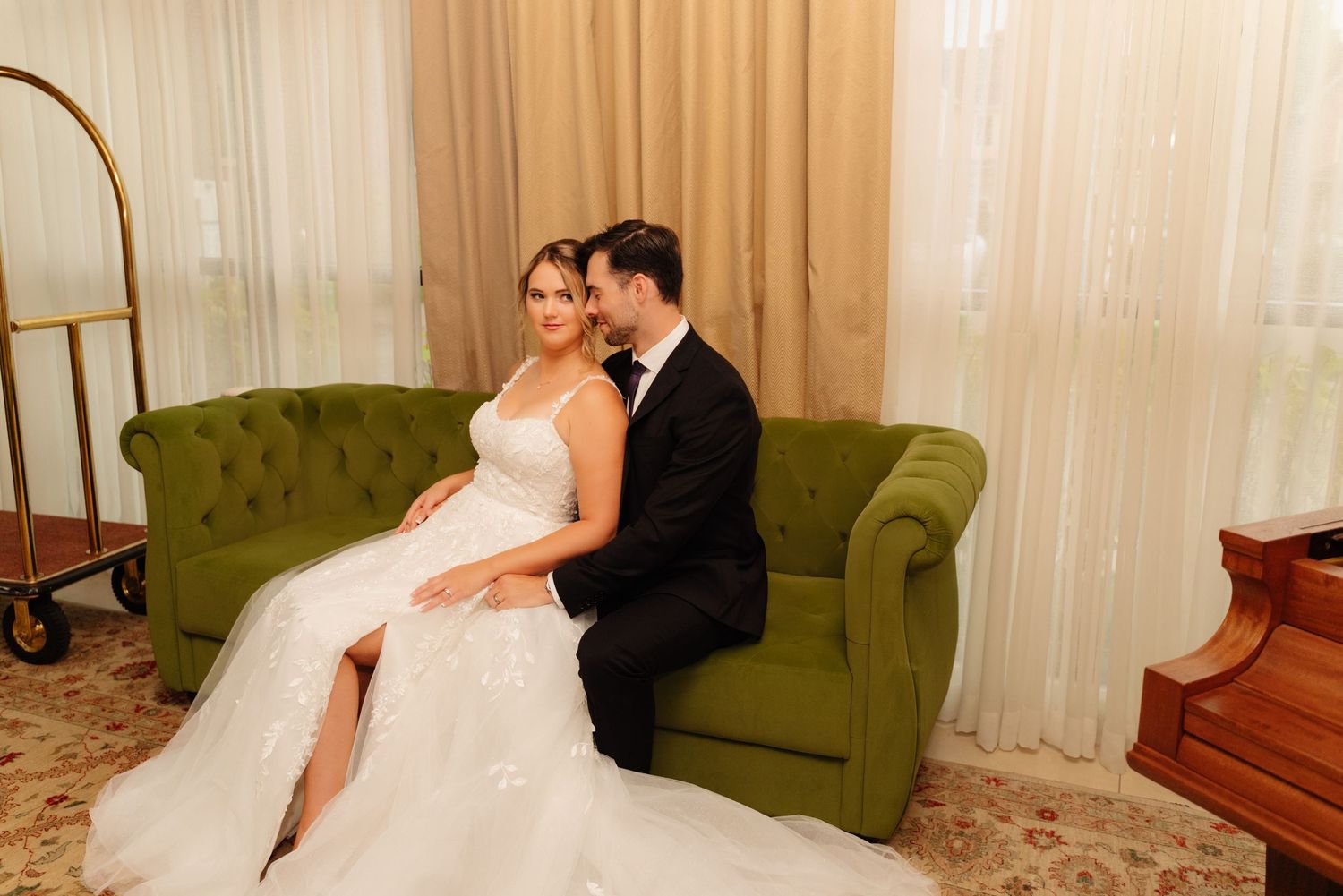 Wedding couple sits together on a green tufted sofa near a grand piano.