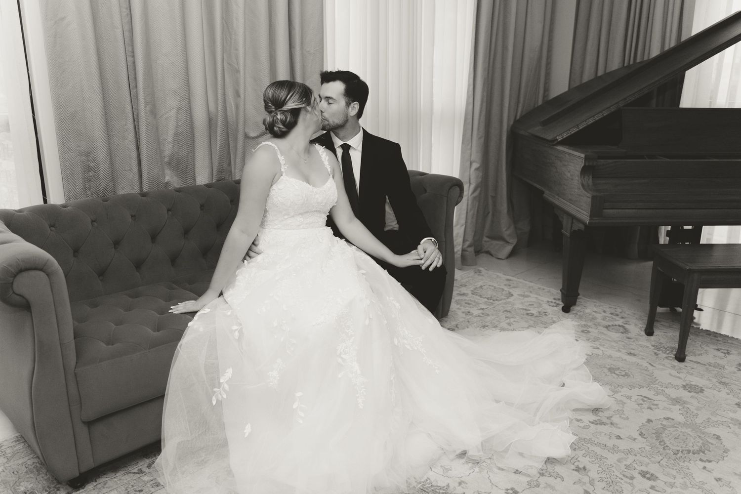 Black and white portrait of newlyweds on vintage sofa with piano in elegant setting.