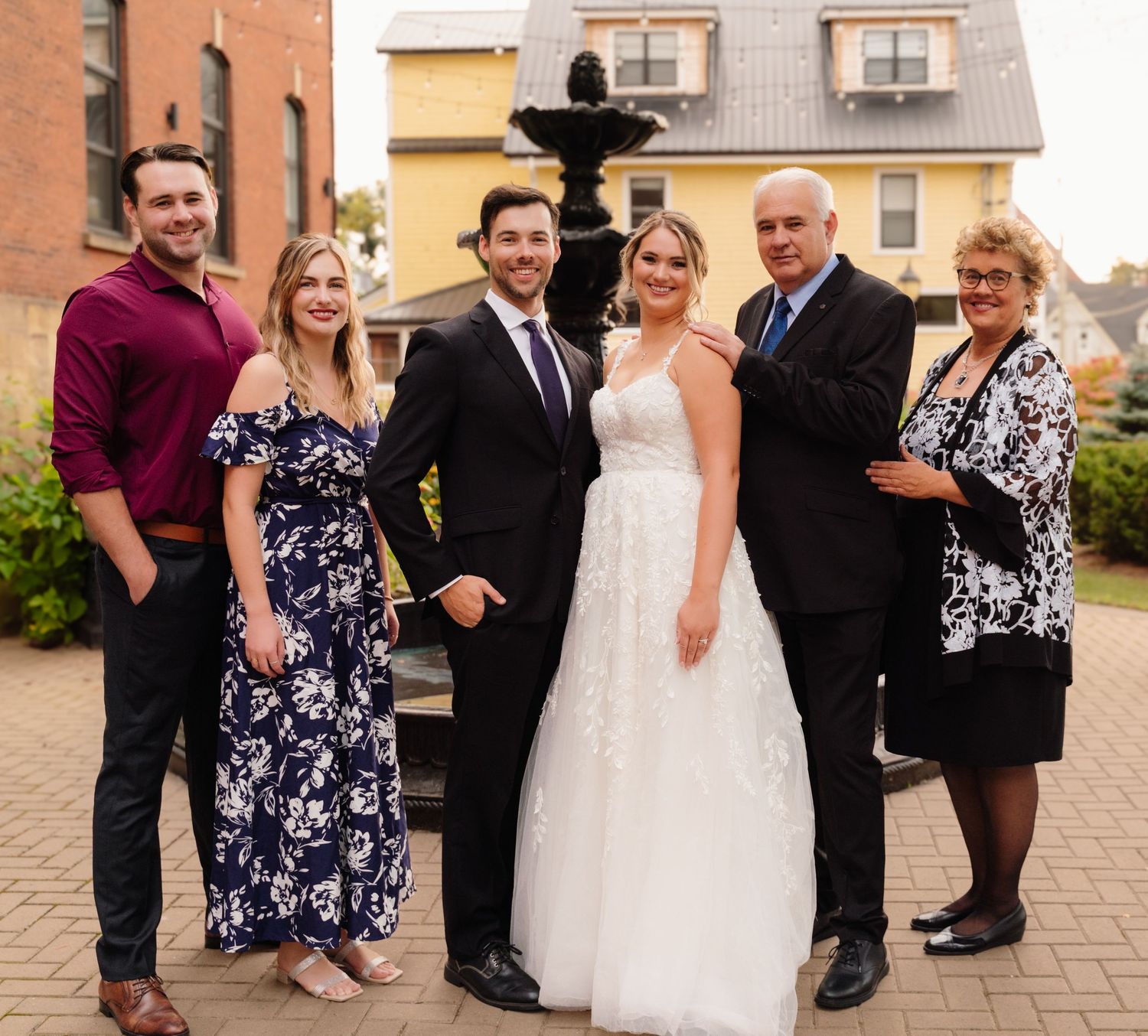 Wedding party in formal attire poses together by fountain against brick and yellow building backdrop.