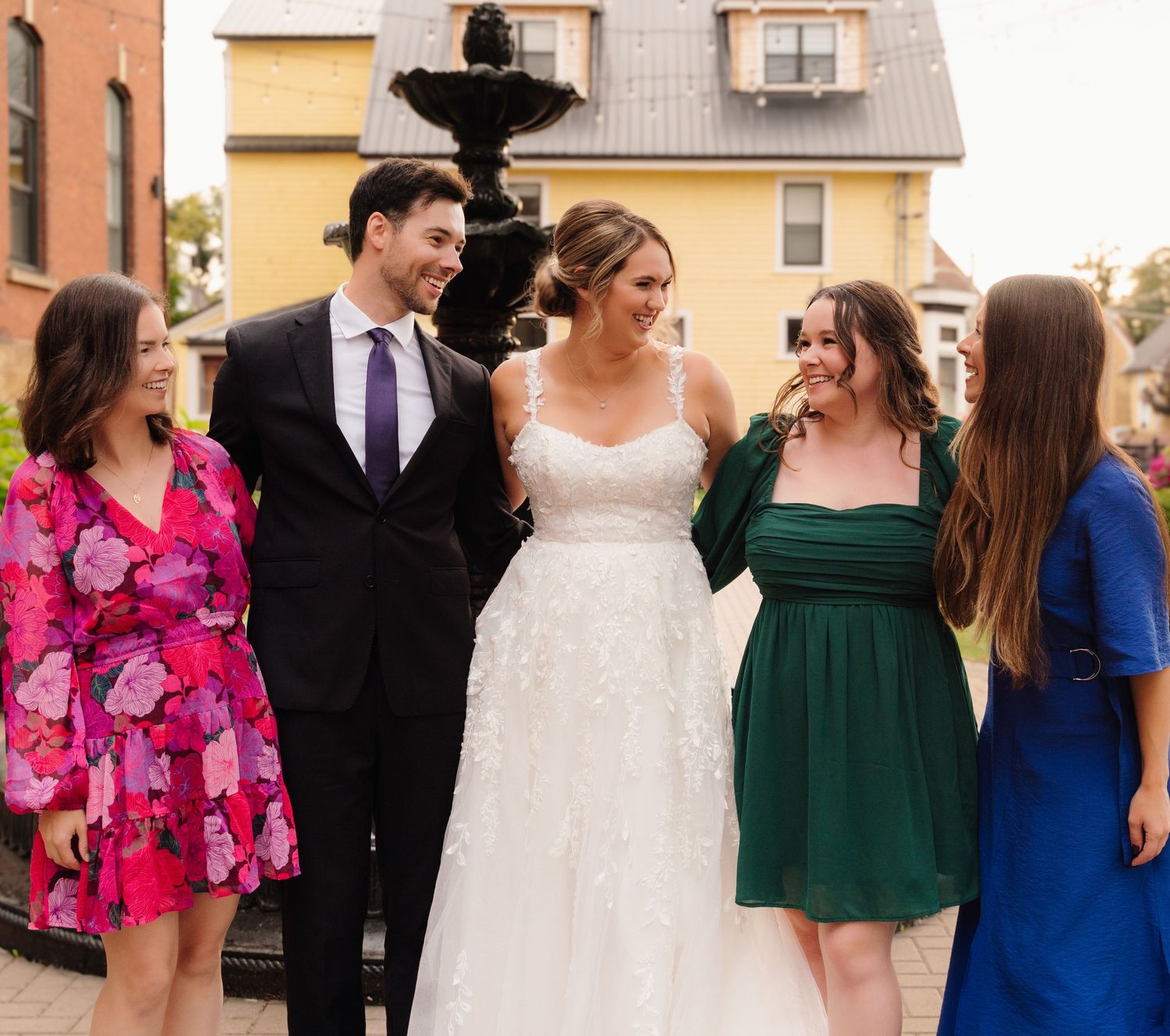 Wedding group in mix of formal attire poses near decorative outdoor fountain.