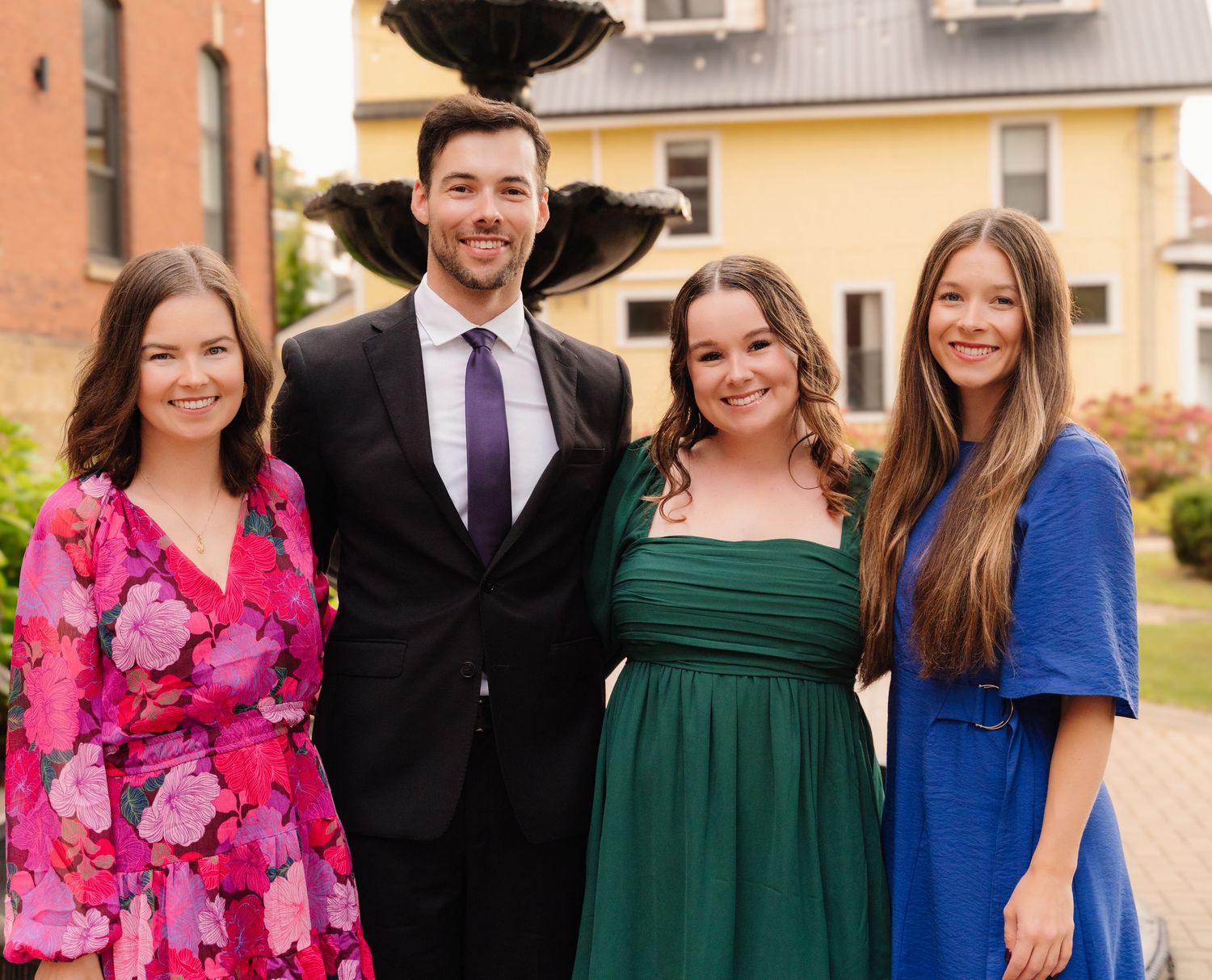 Four people in vibrant formal wear smile near tiered fountain and yellow building.