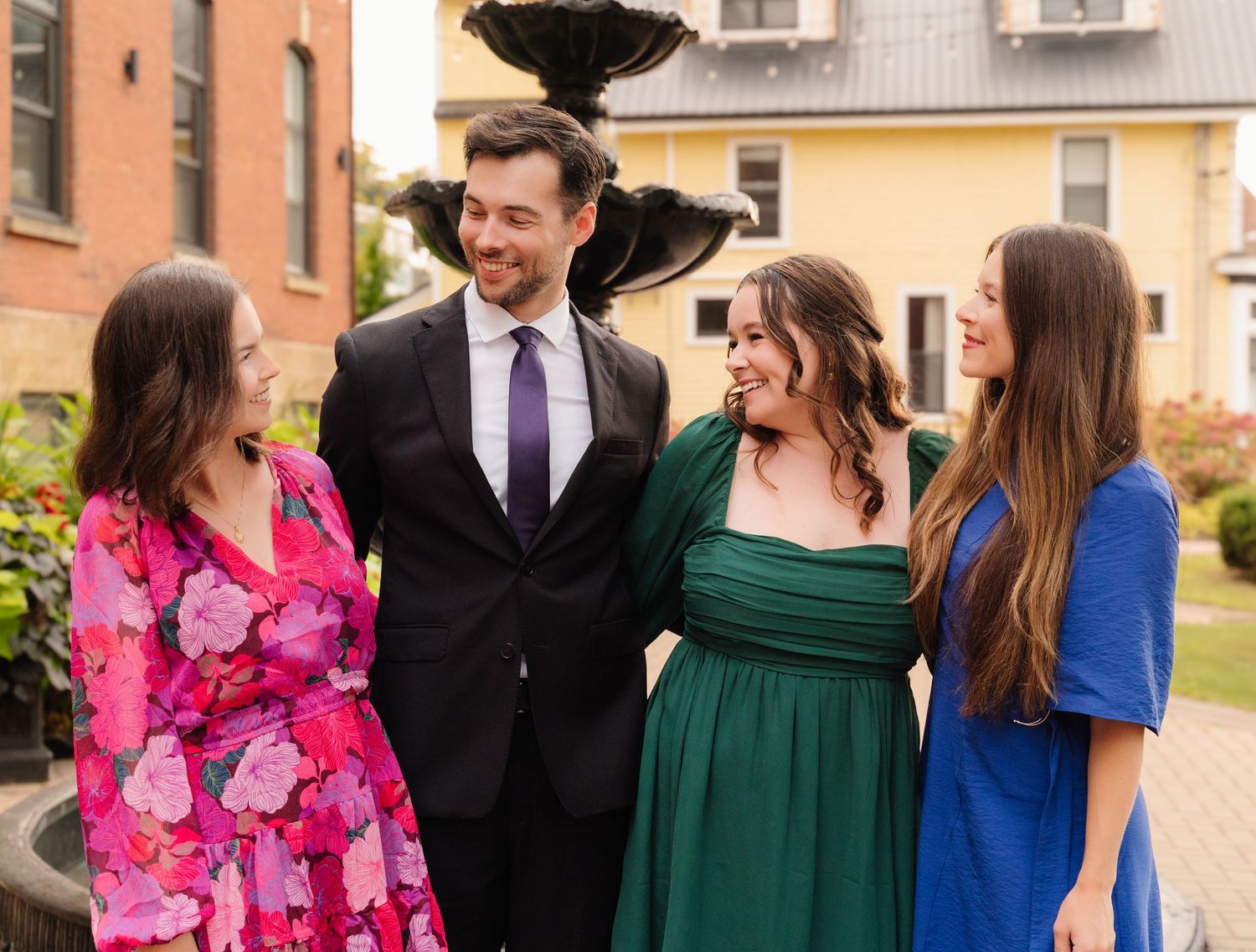 Group of four in colorful dresses and suit chat casually by outdoor fountain.