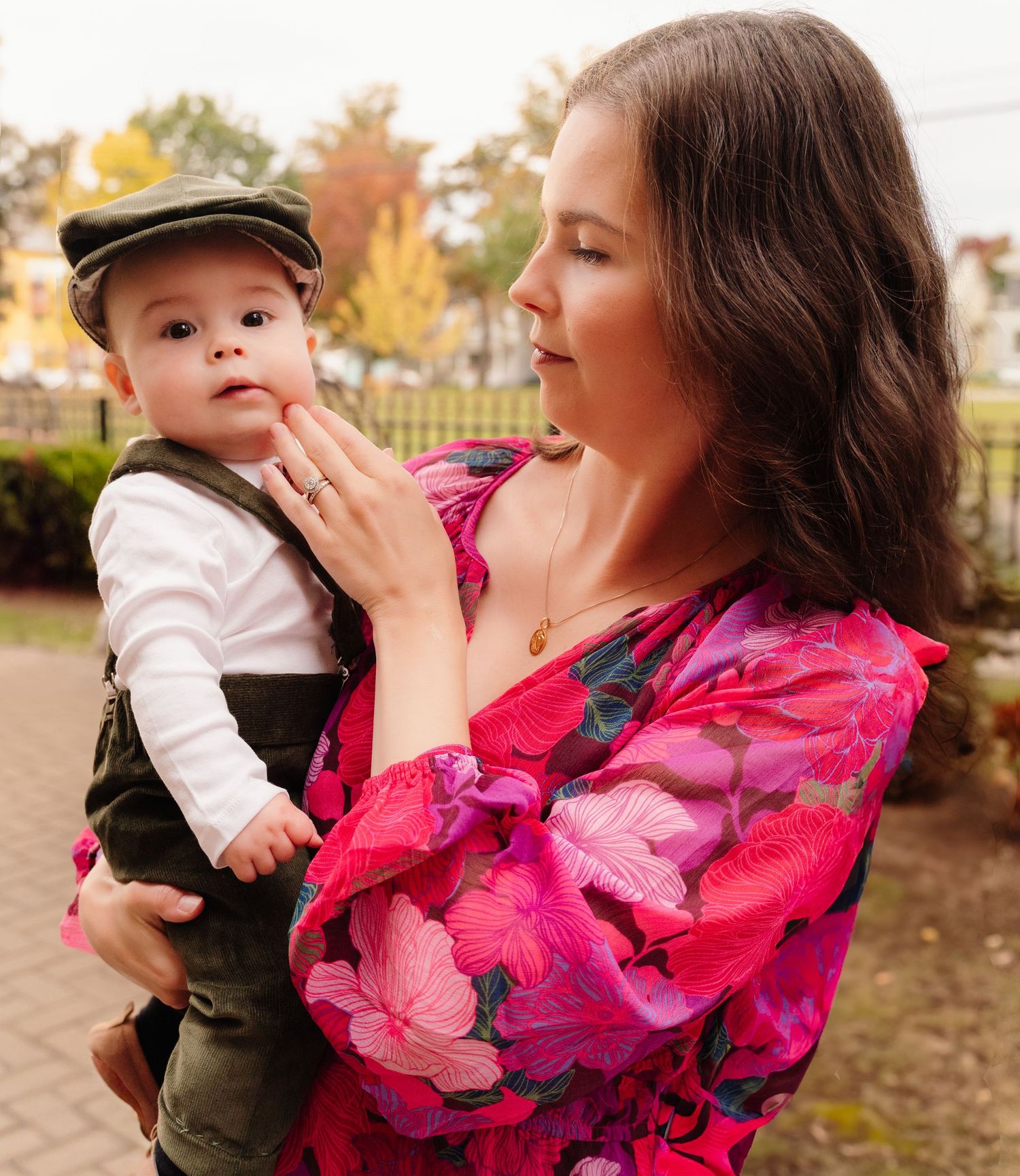 Person in floral dress holds baby wearing newsboy cap outdoors in fall setting.