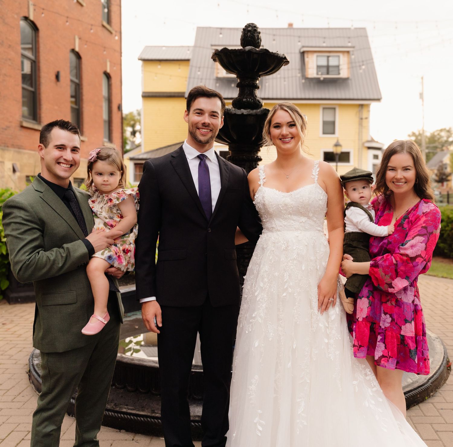 Wedding couple with guests pose by black fountain against brick building backdrop.