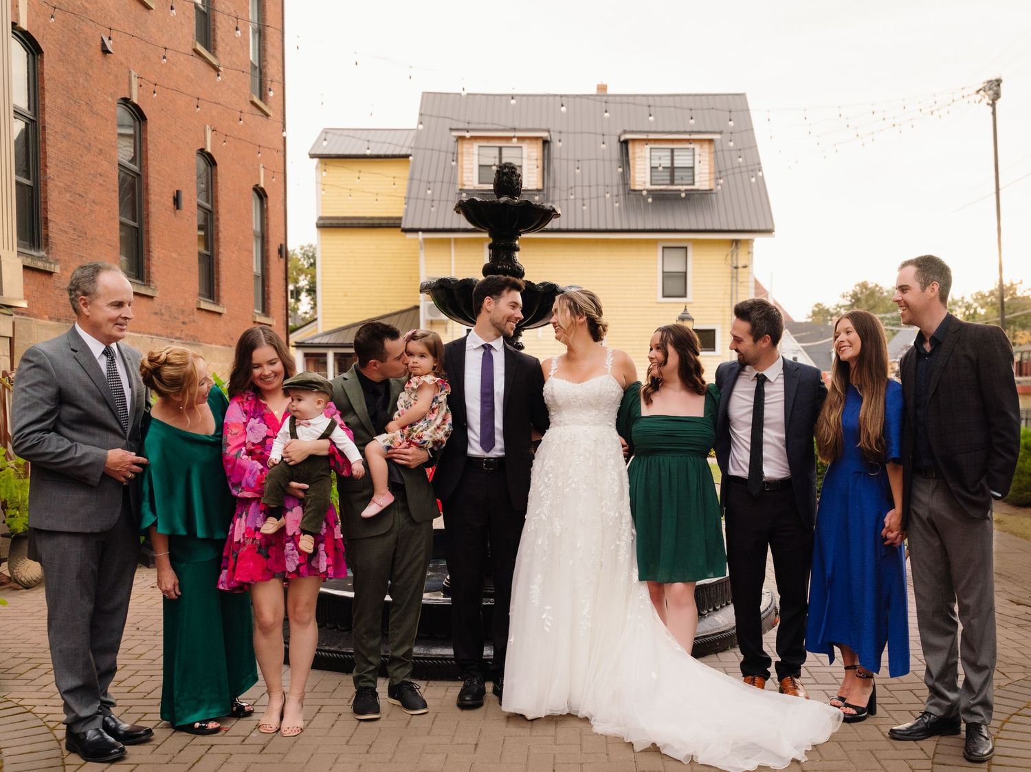 Wedding party poses outdoors by a brick building with decorative fountain and yellow house in background.
