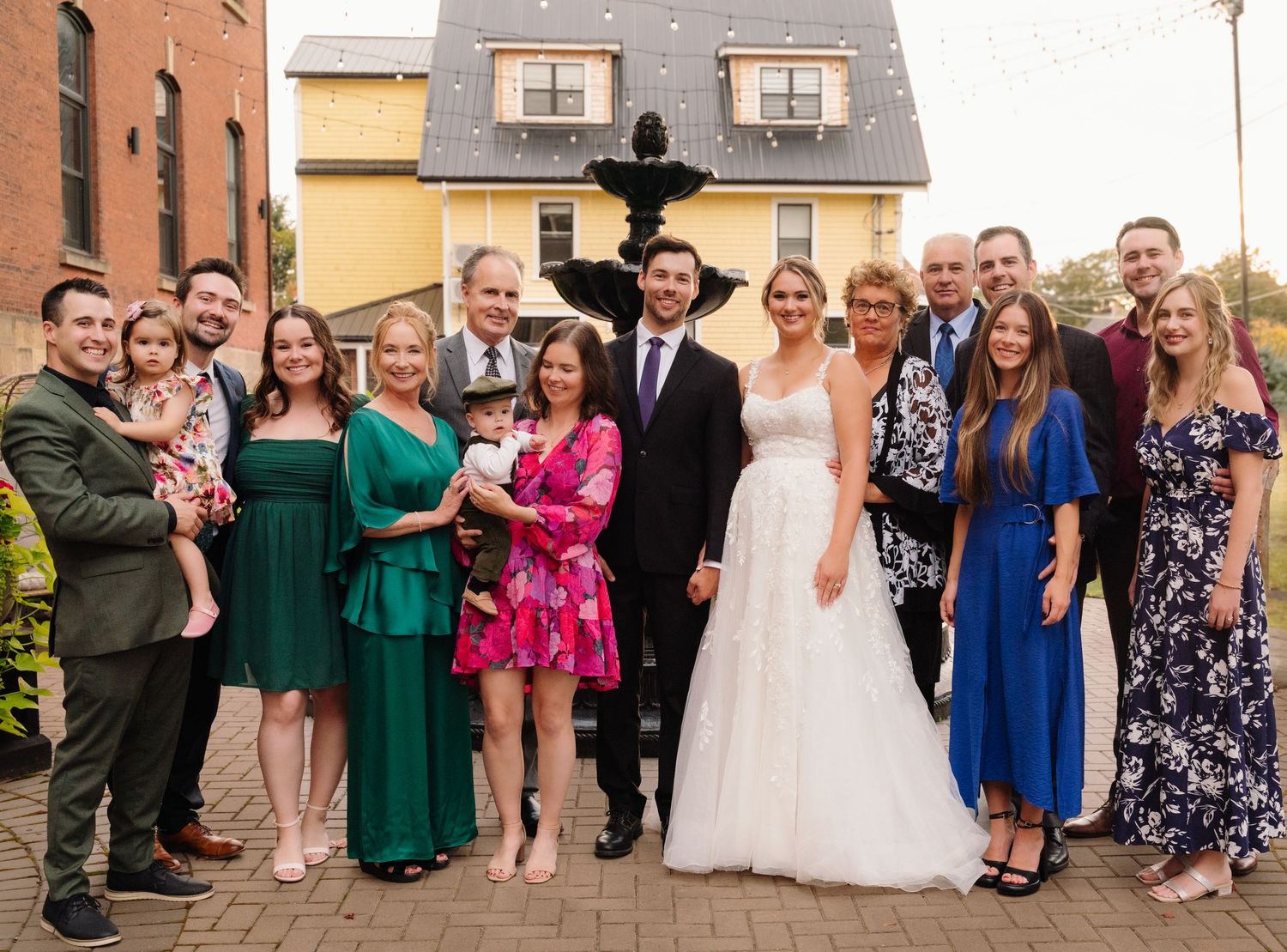 Wedding party gathered for formal photo in front of fountain and brick building.