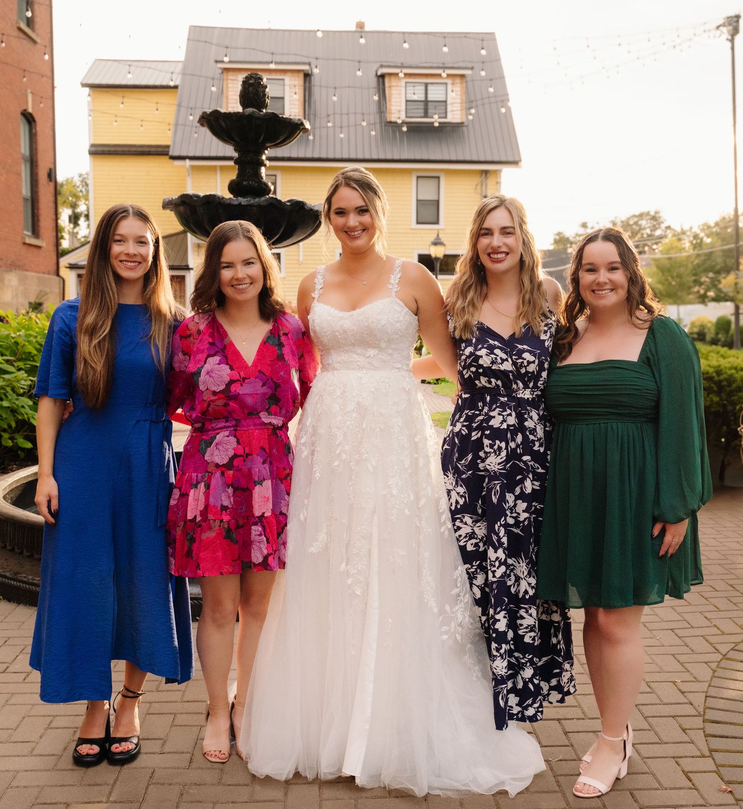 Group of women in colorful dresses pose with bride by decorative fountain.