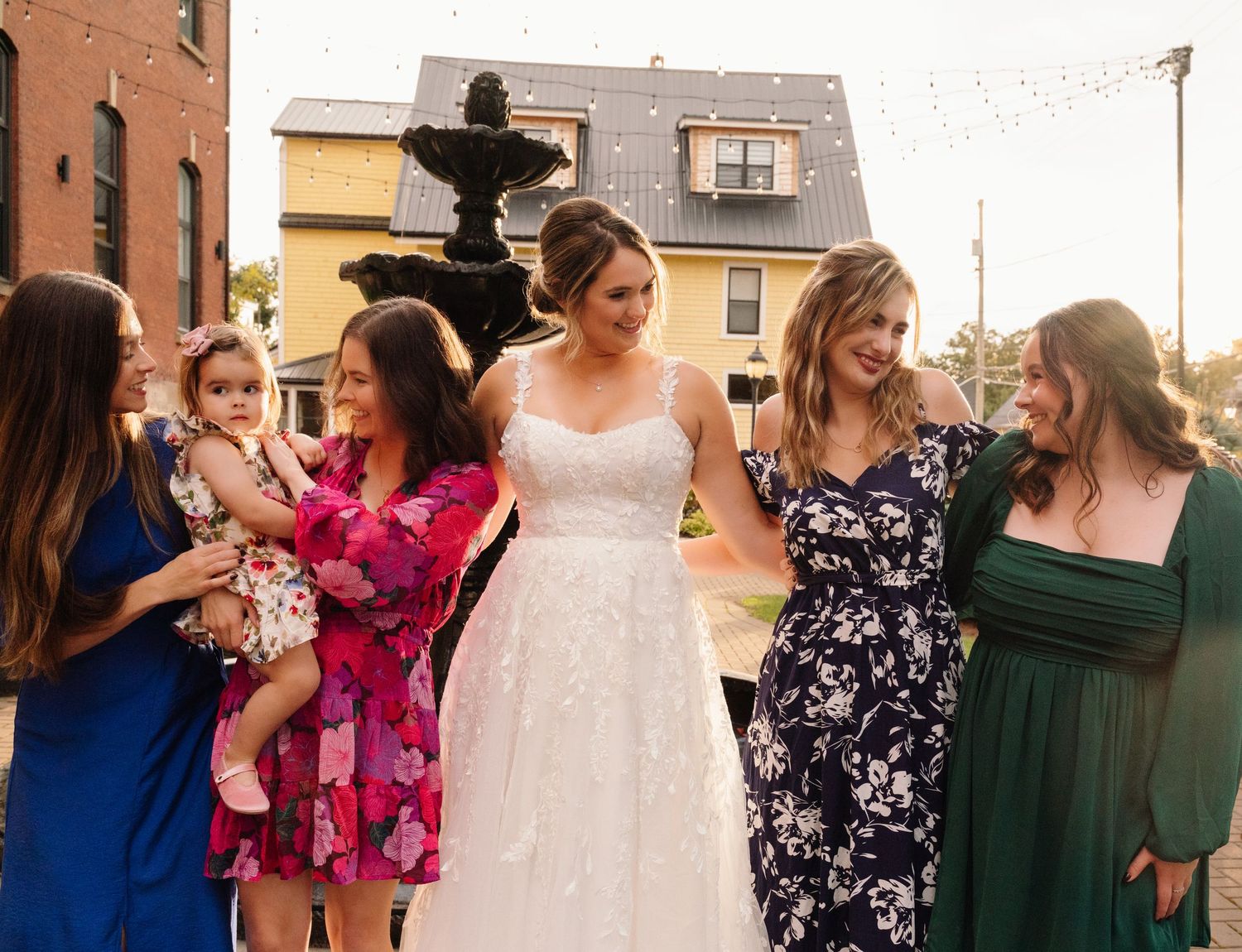 Wedding party members in mix of formal wear gather around bride outdoors.