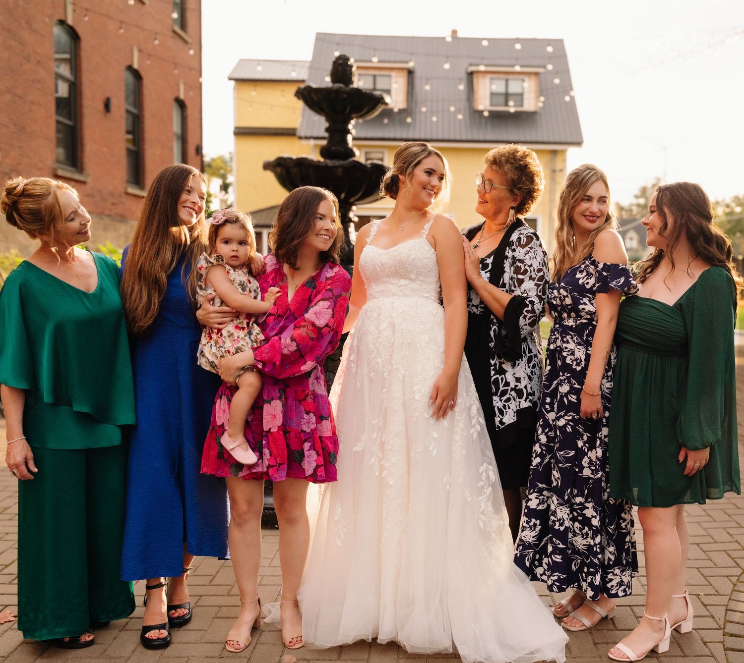 Wedding party group stands by ornate fountain with guests in colorful dresses against brick building backdrop.