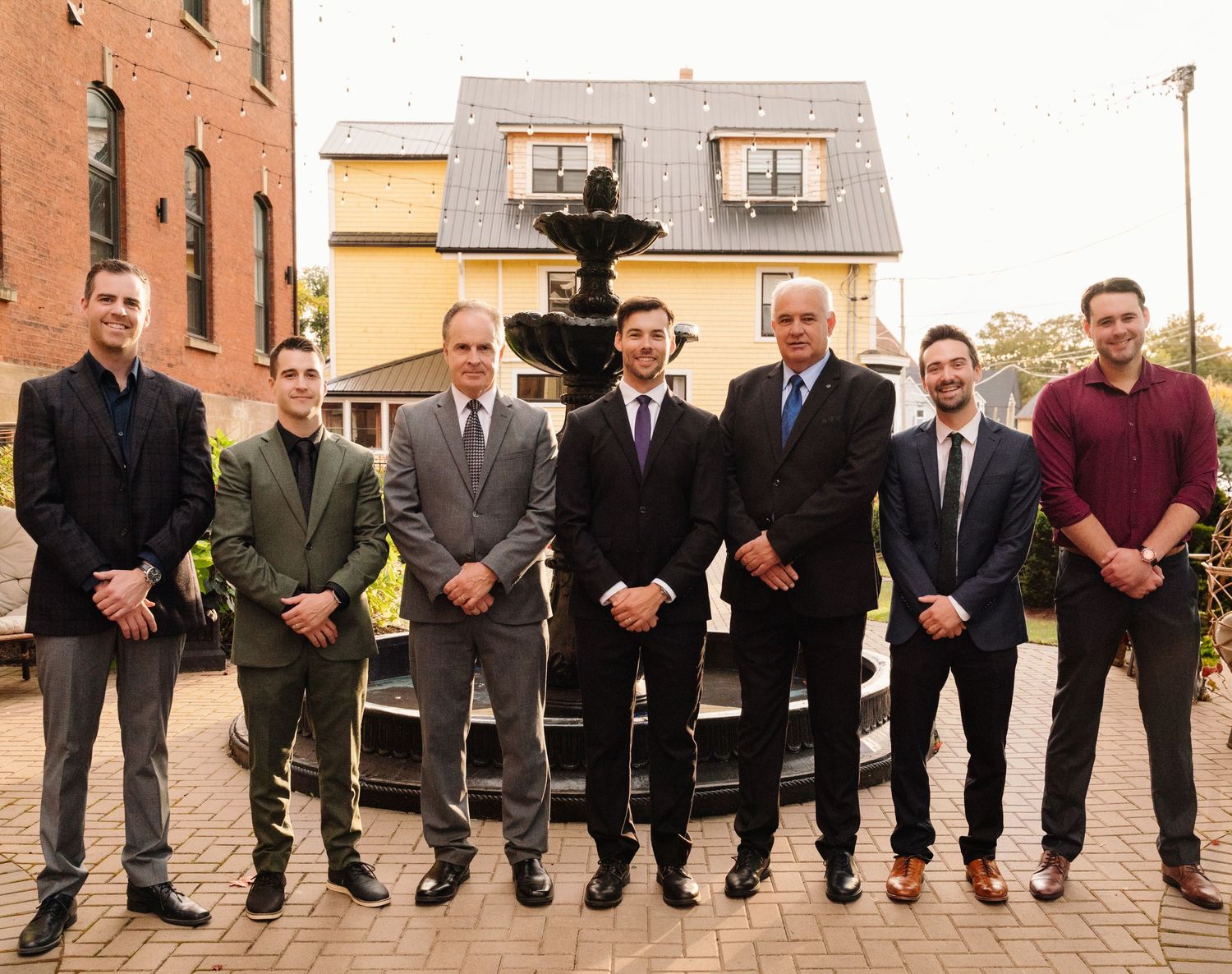 Groomsmen in formal attire pose together in front of decorative fountain and brick building.
