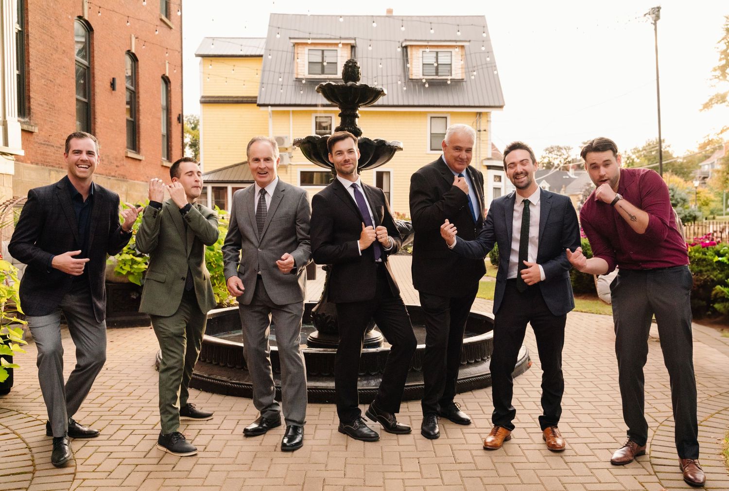 Group of men in suits making playful poses by fountain during wedding celebration.