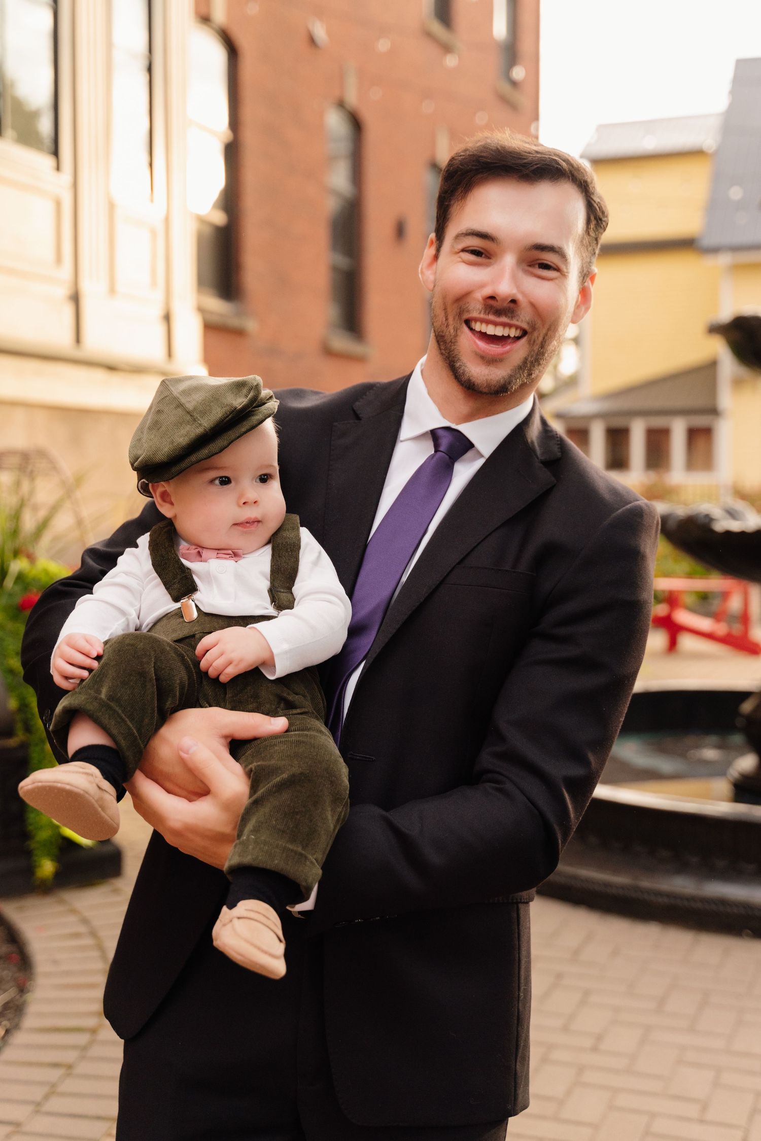 Formal-dressed person holds small child wearing vintage-style overalls and cap outdoors.