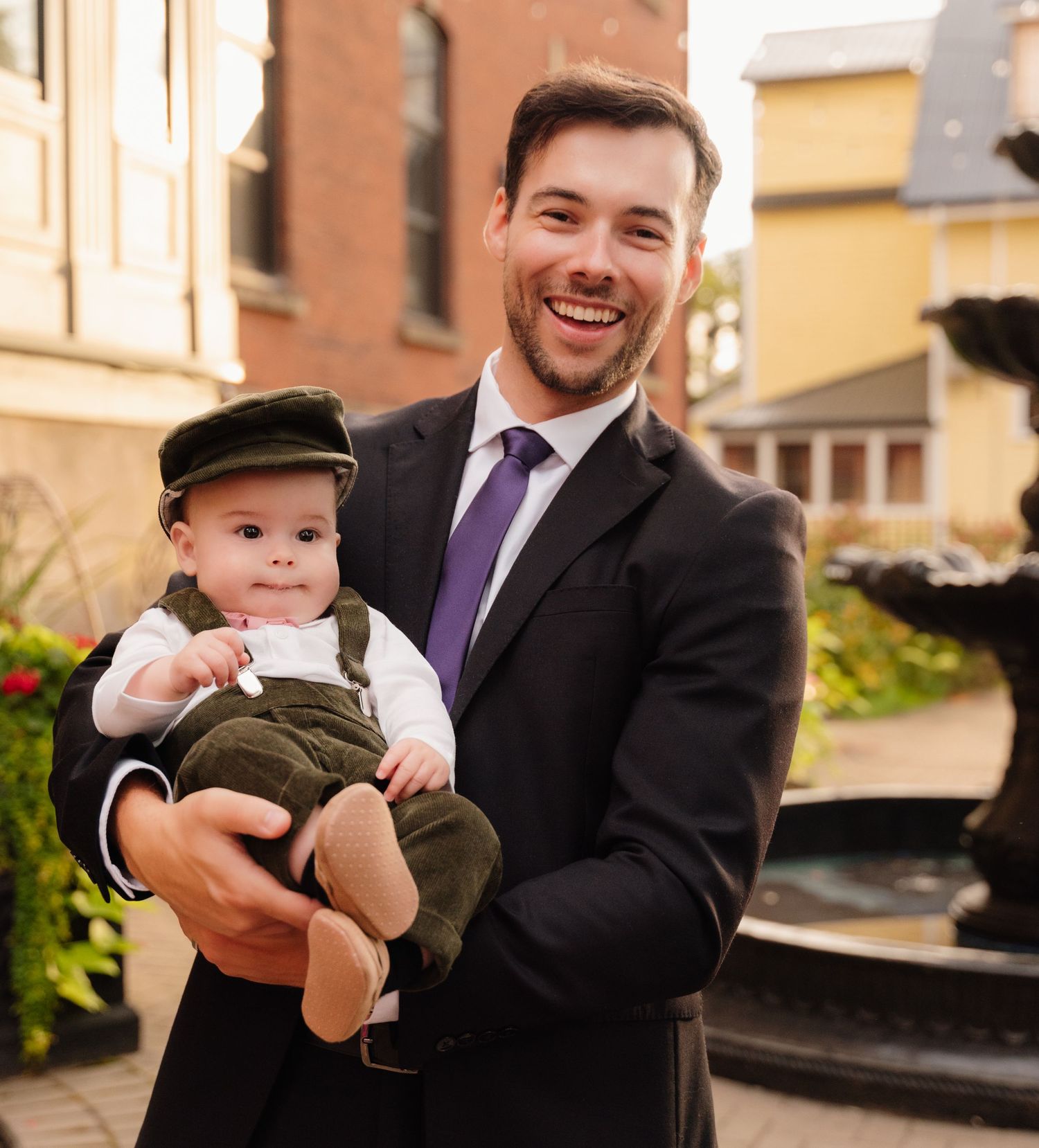 Person in suit holds smiling toddler wearing olive green overalls and cap by brick building.
