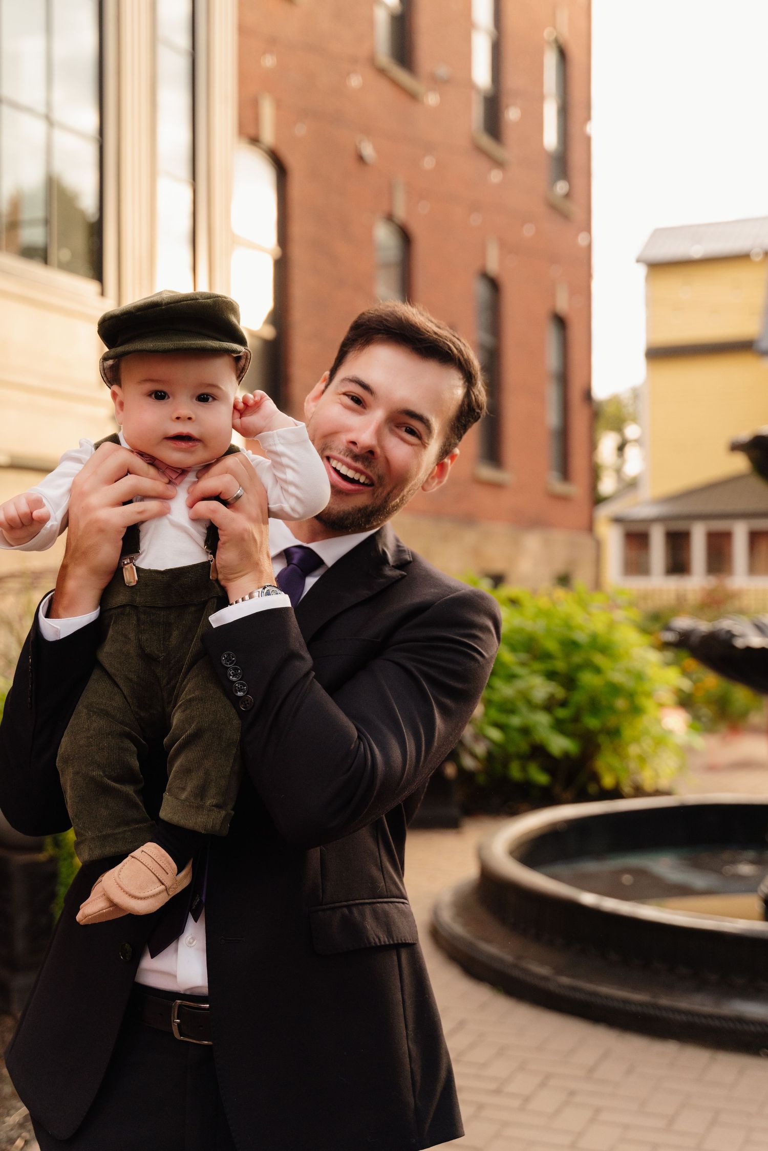 Person in dark suit holds baby wearing vintage-style outfit near decorative fountain.