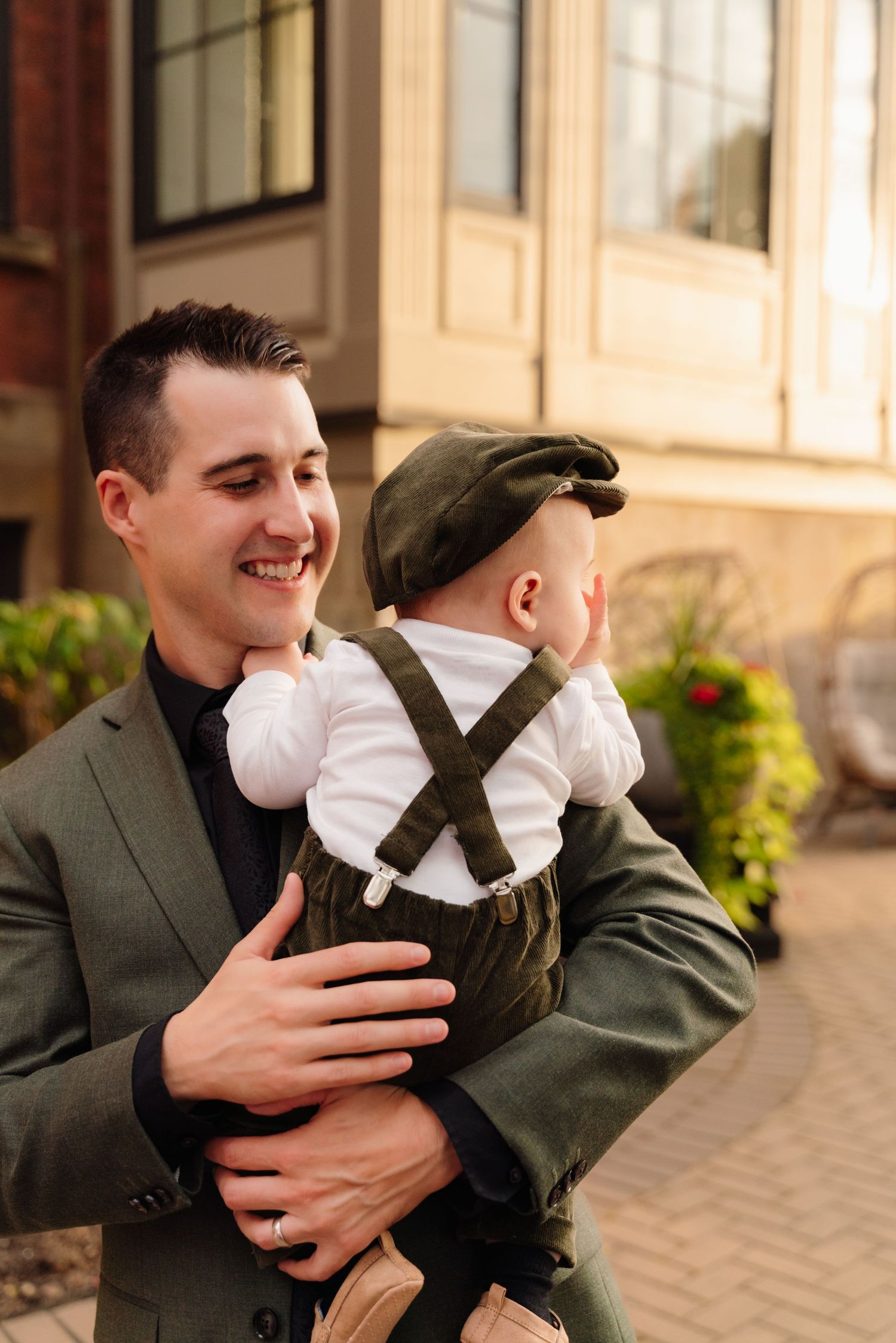 Person in grey suit holds infant wearing white shirt and olive green overalls outdoors.