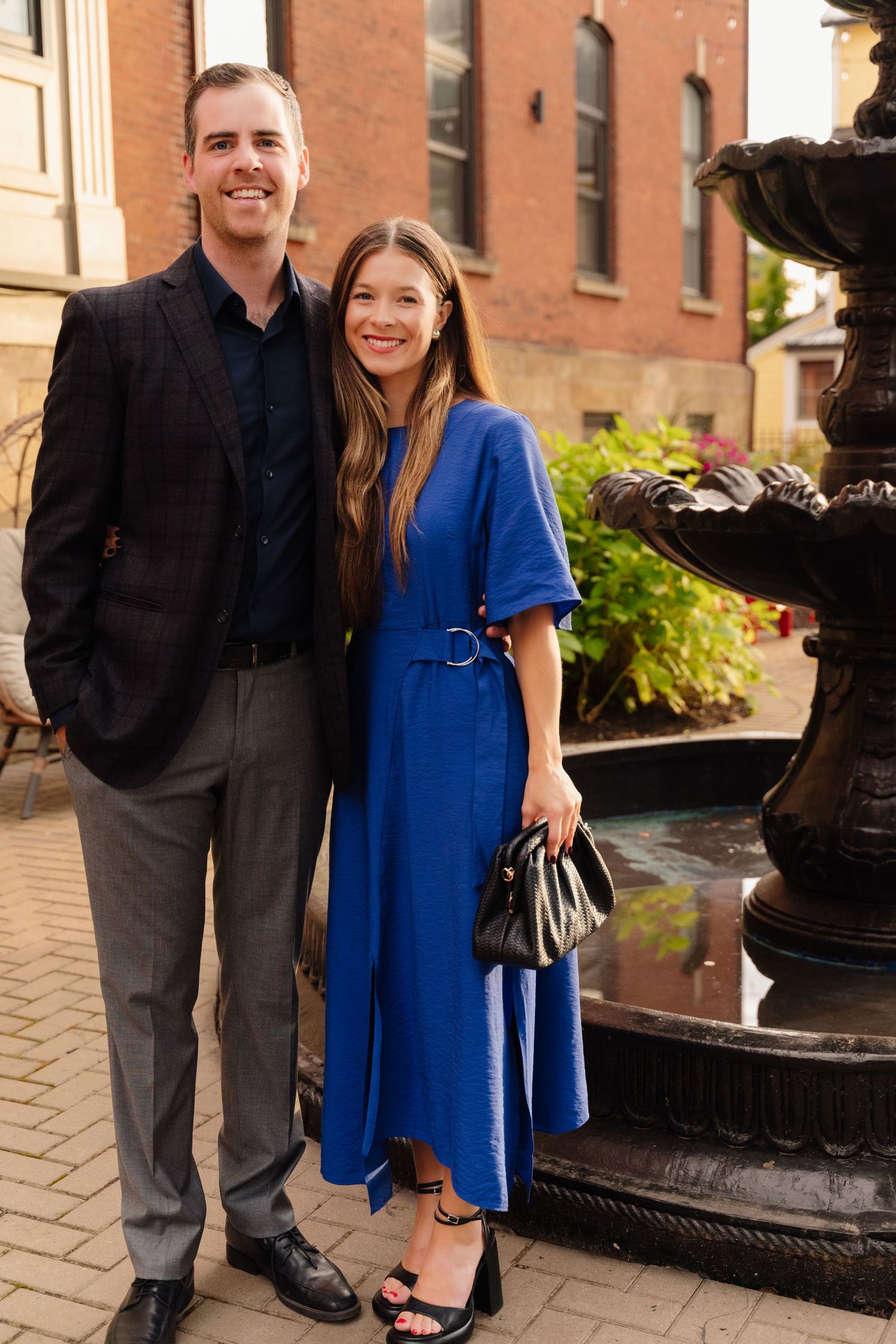 Couple in formal wear poses together near ornate fountain and brick building.