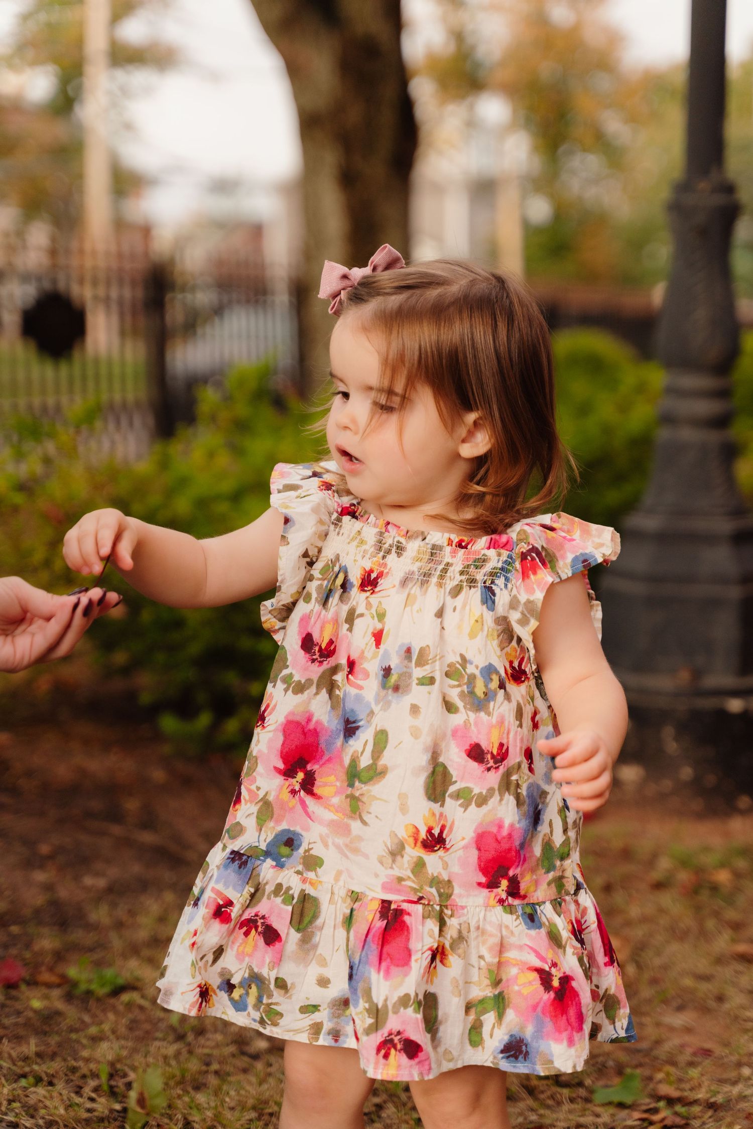 Small child in floral dress reaches out while playing outdoors.