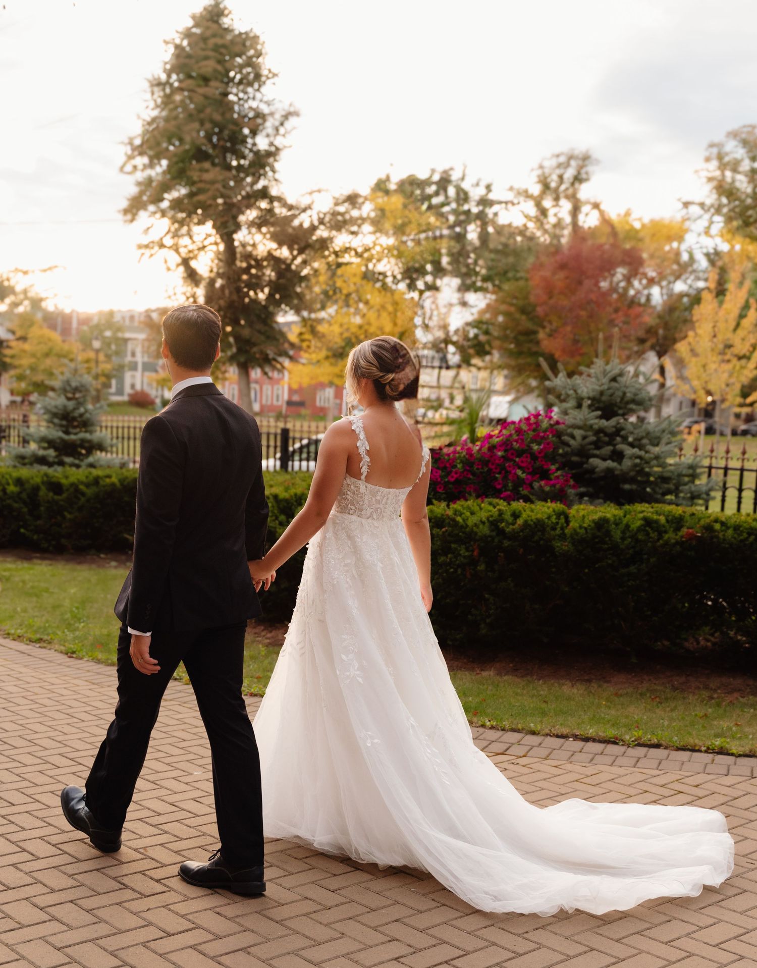 A newlywed couple walks along a brick path surrounded by autumn foliage.