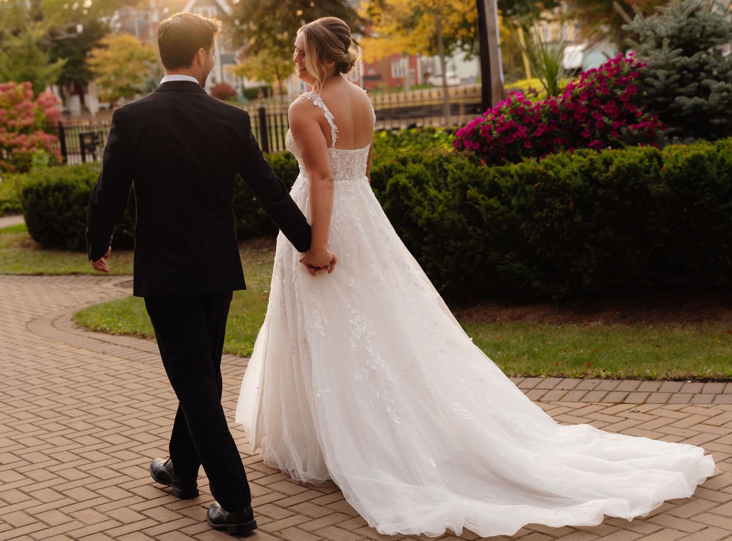 A wedding couple strolls hand in hand on a brick walkway with fall colors in the background.
