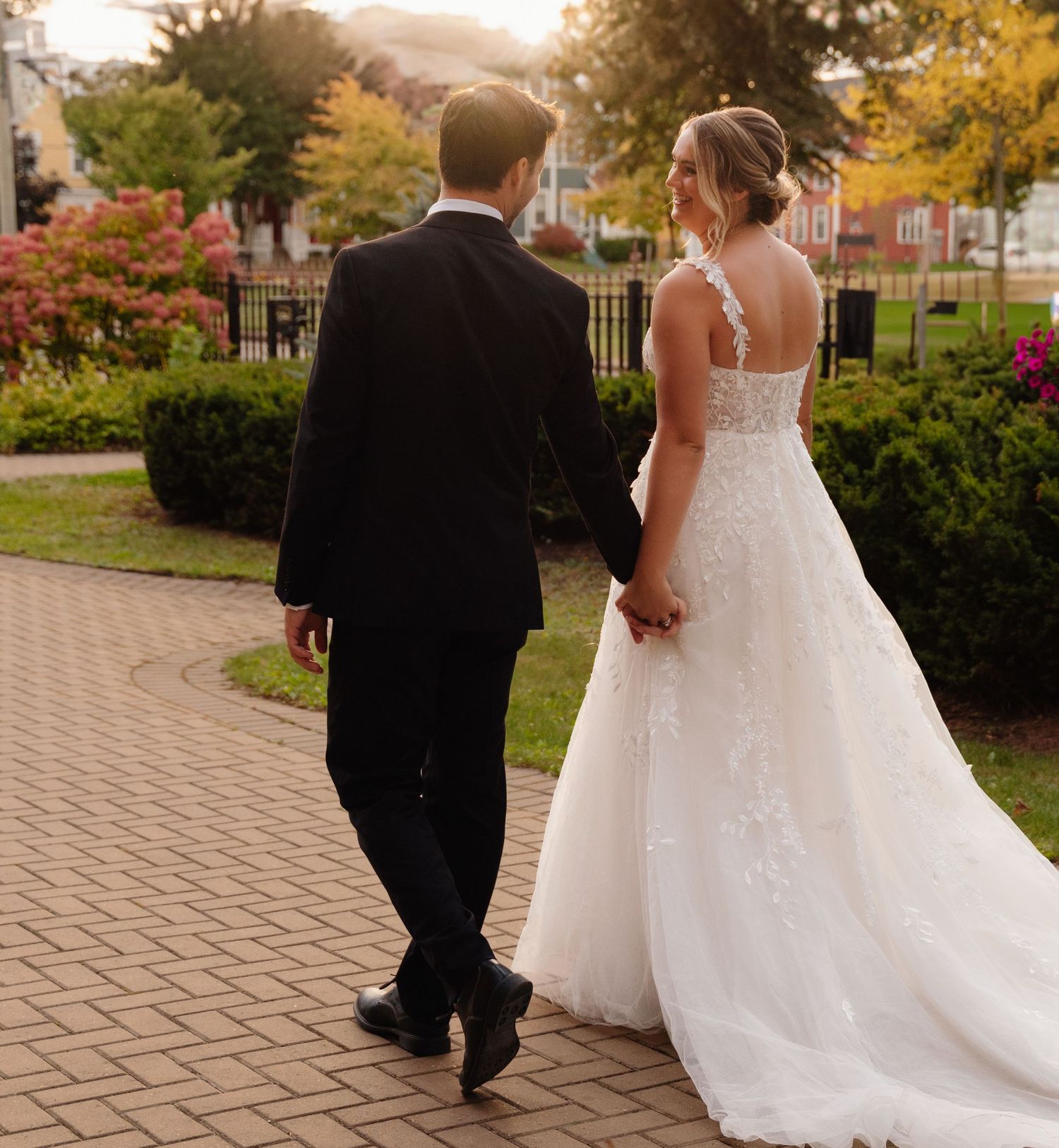 Newlyweds walk together on a brick path during golden hour with autumn trees behind them.