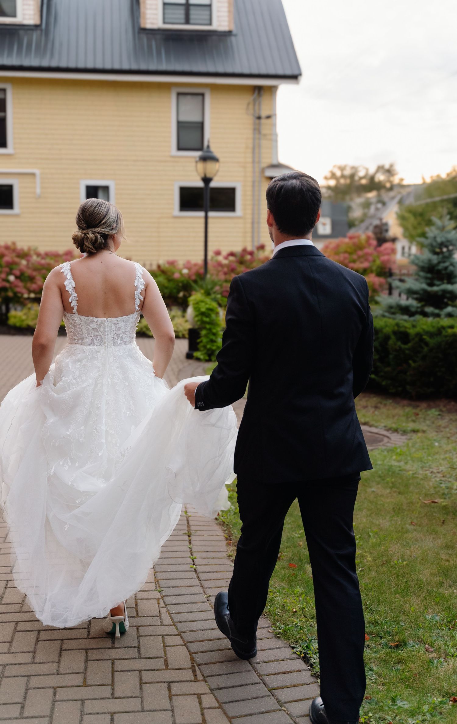 Couple walking on brick pathway with elegant white wedding dress and black suit near yellow house.