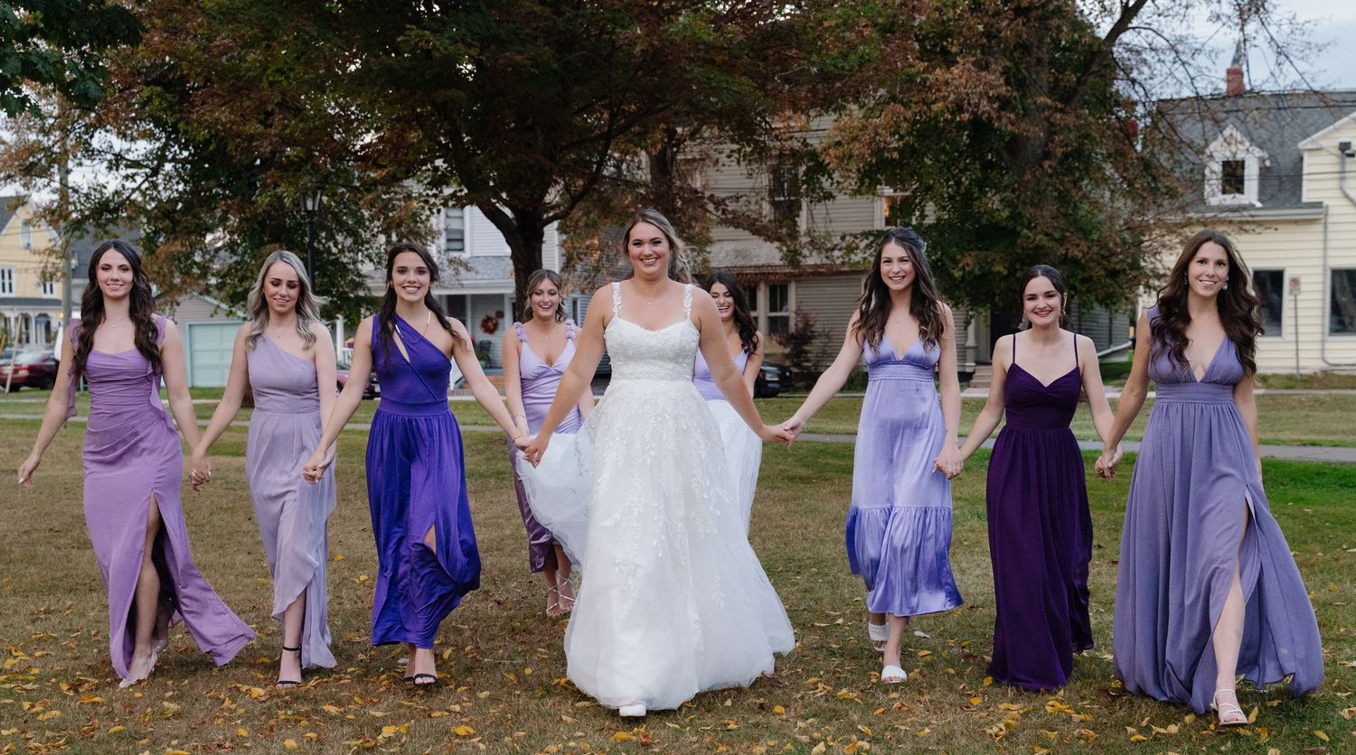 Bride and bridesmaids walk together wearing coordinating purple and lavender dresses in fall setting.