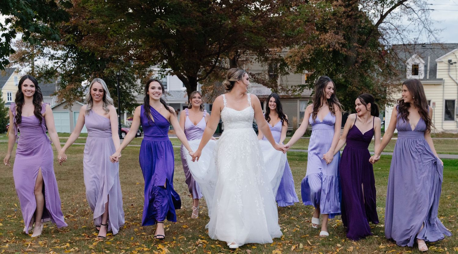 Bridal party in shades of purple dresses poses together on autumn lawn.