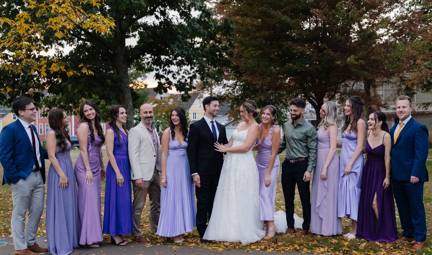 Large wedding party in purple and blue attire poses together on fallen autumn leaves.
