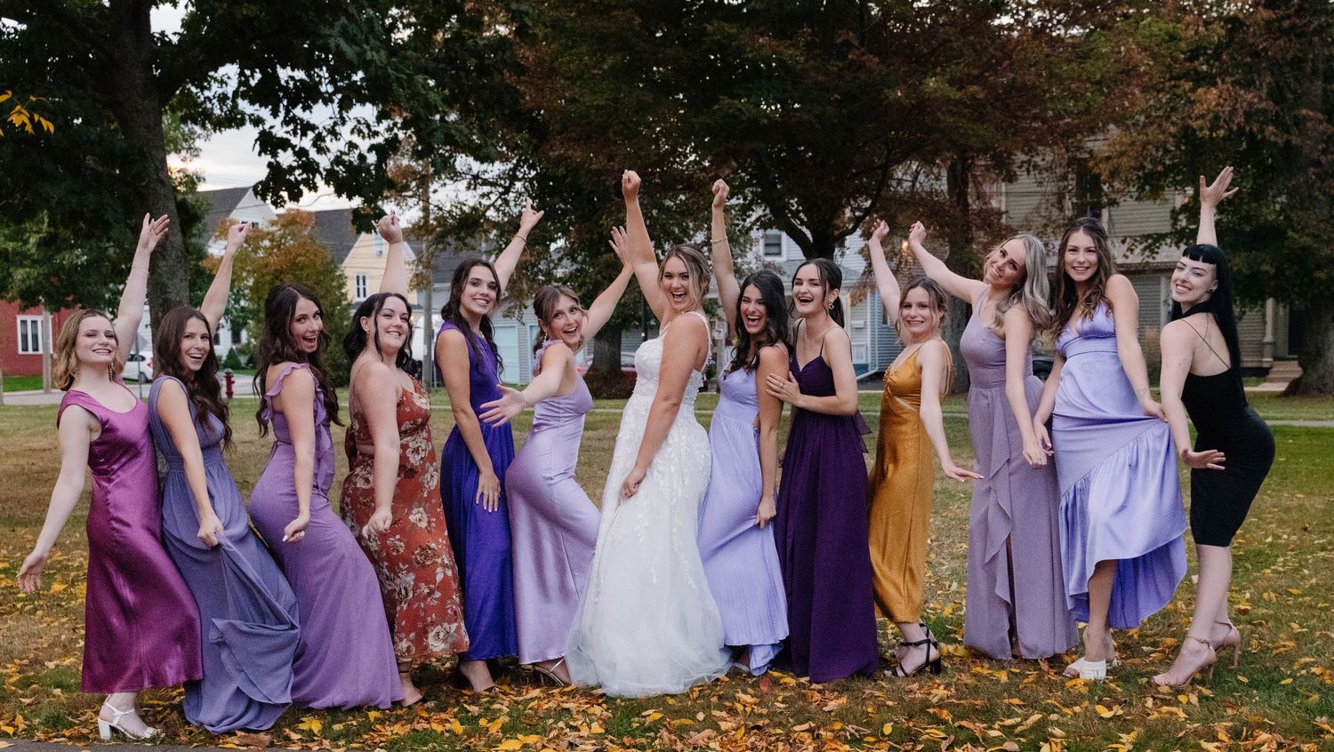 Wedding party in purple and lavender dresses celebrating with raised hands among autumn leaves.