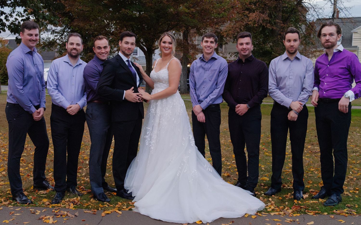 Wedding party with groomsmen in matching attire surrounding couple on autumn day.