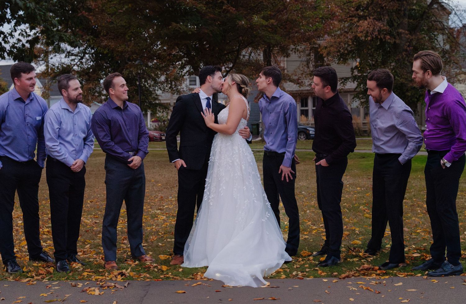 Wedding couple and groomsmen posing together on leaf-covered ground in fall setting.