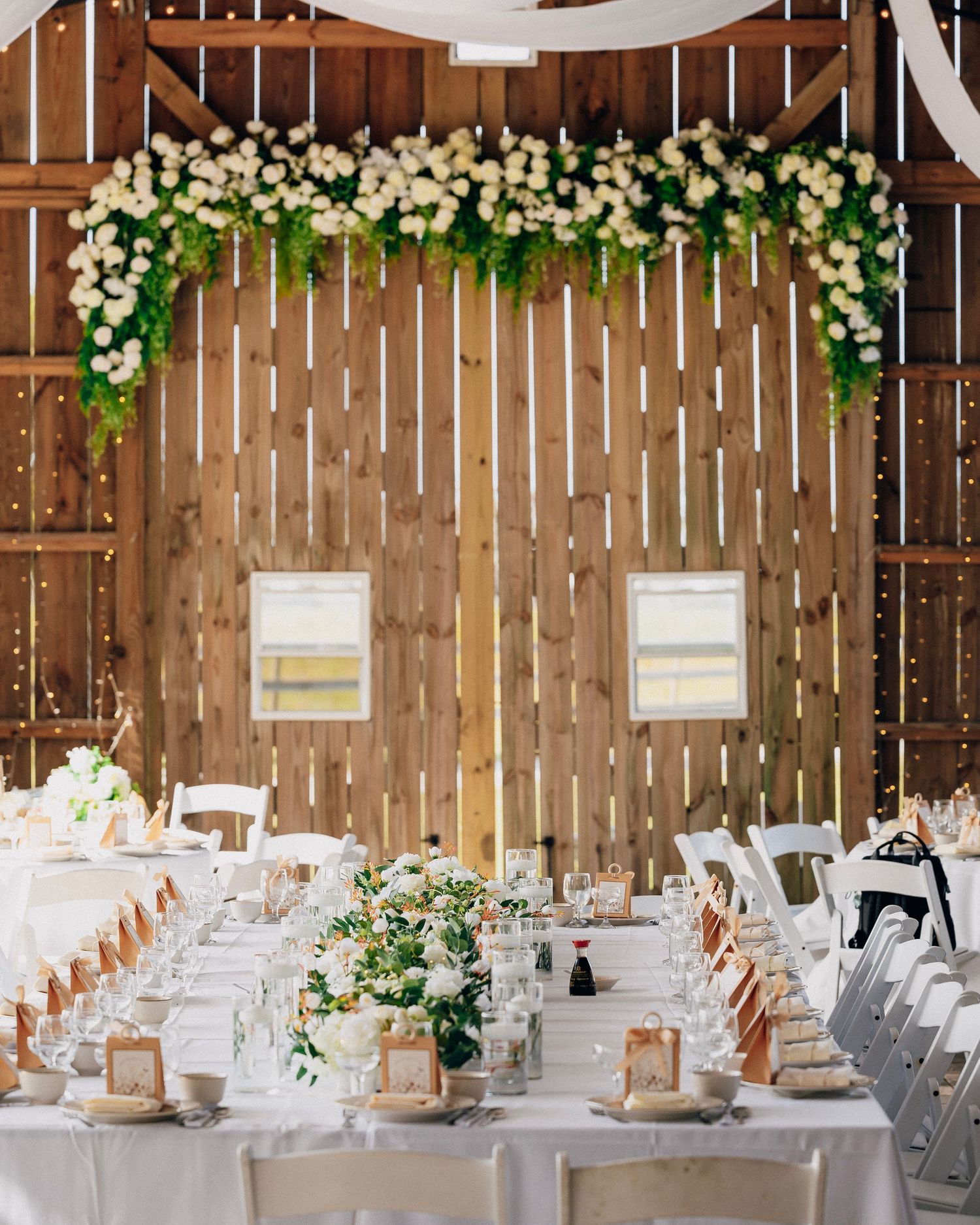Elegant barn wedding reception with long white tables, floral arch of white roses and greenery on rustic wood wall.