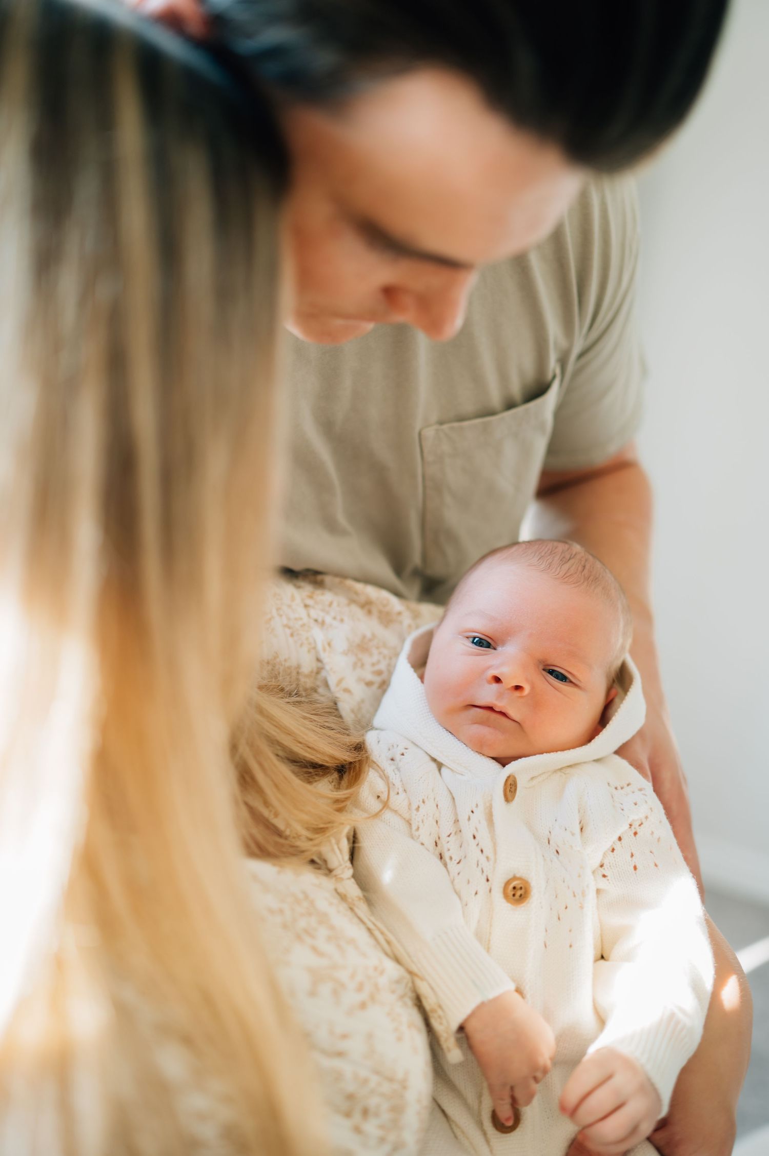 Parents snuggling with newborn baby wearing white cardigan in sunlit room.