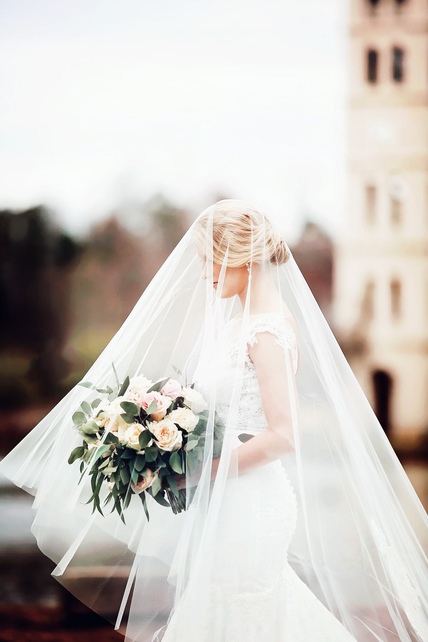 Bride in flowing veil holds elegant white and green bouquet against church backdrop.