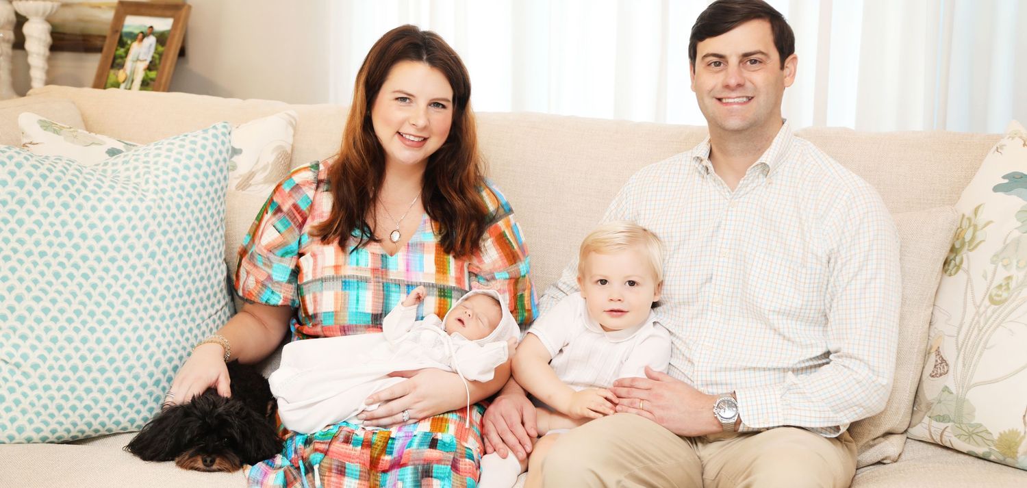A family sits together on a couch with their newborn baby and a black cat in a bright living room.