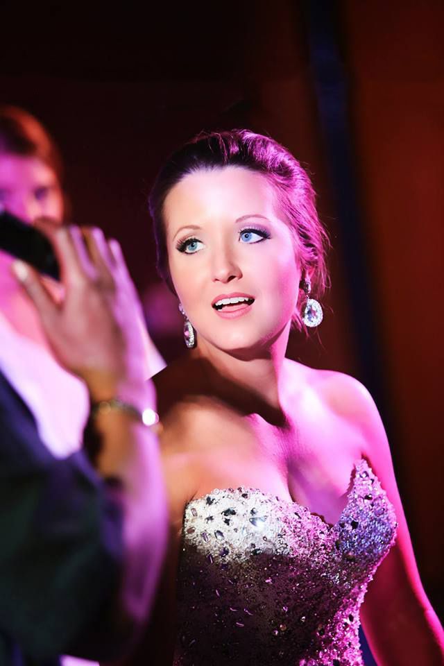 Performer in a sparkly strapless gown sings on stage under purple lighting.