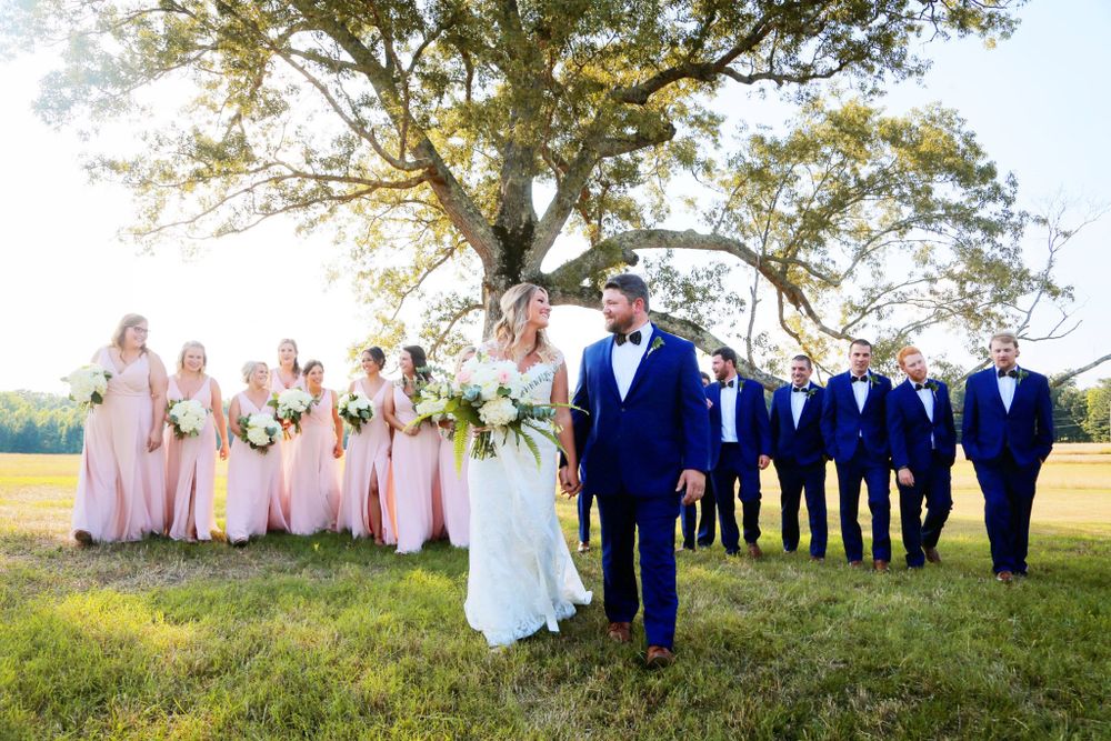 A wedding party in pink and blue attire poses under a large tree in a sunny field.