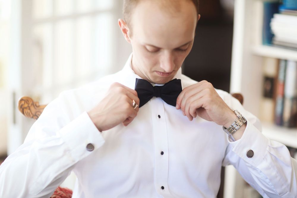 A person adjusts their black bow tie while wearing a white tuxedo shirt.