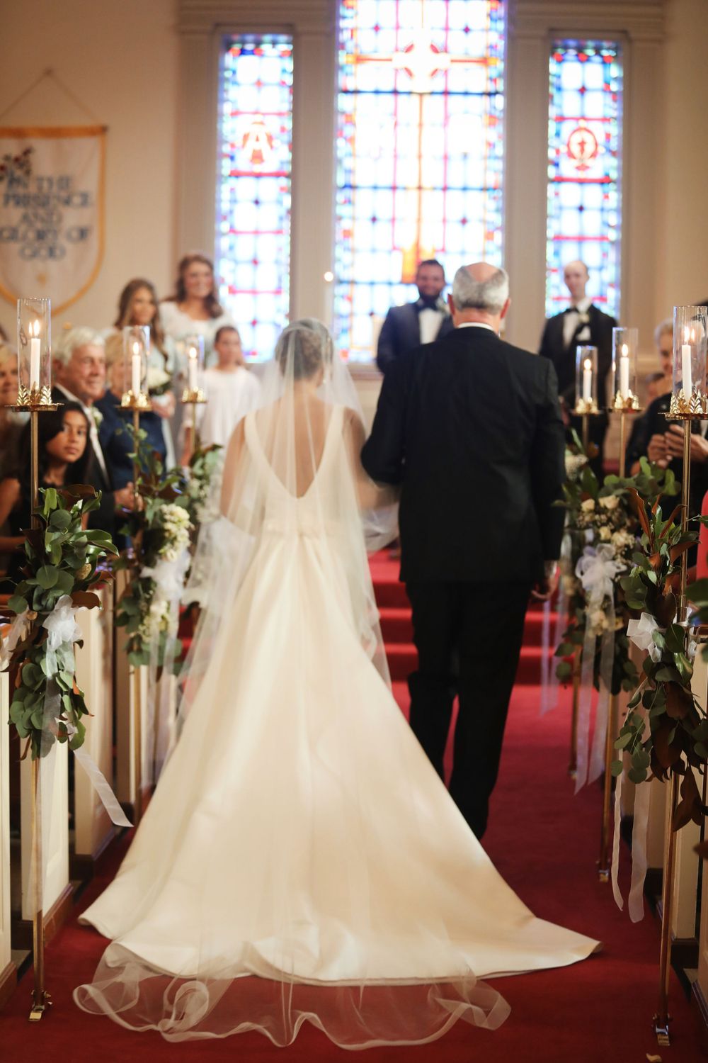 A bride walks down the church aisle with beautiful stained glass windows and floral decorations.