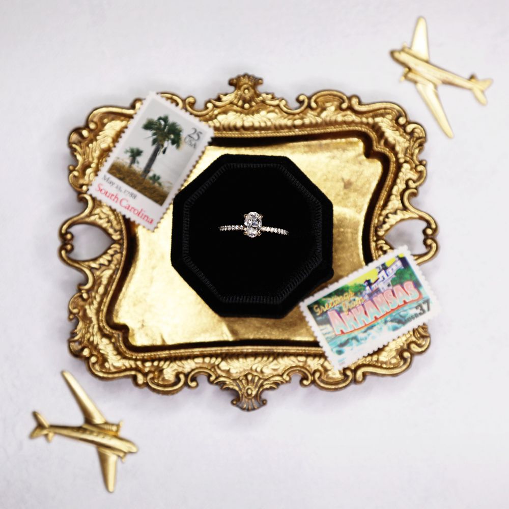 A diamond engagement ring sits in black box on vintage gold tray with miniature airplane decorations.