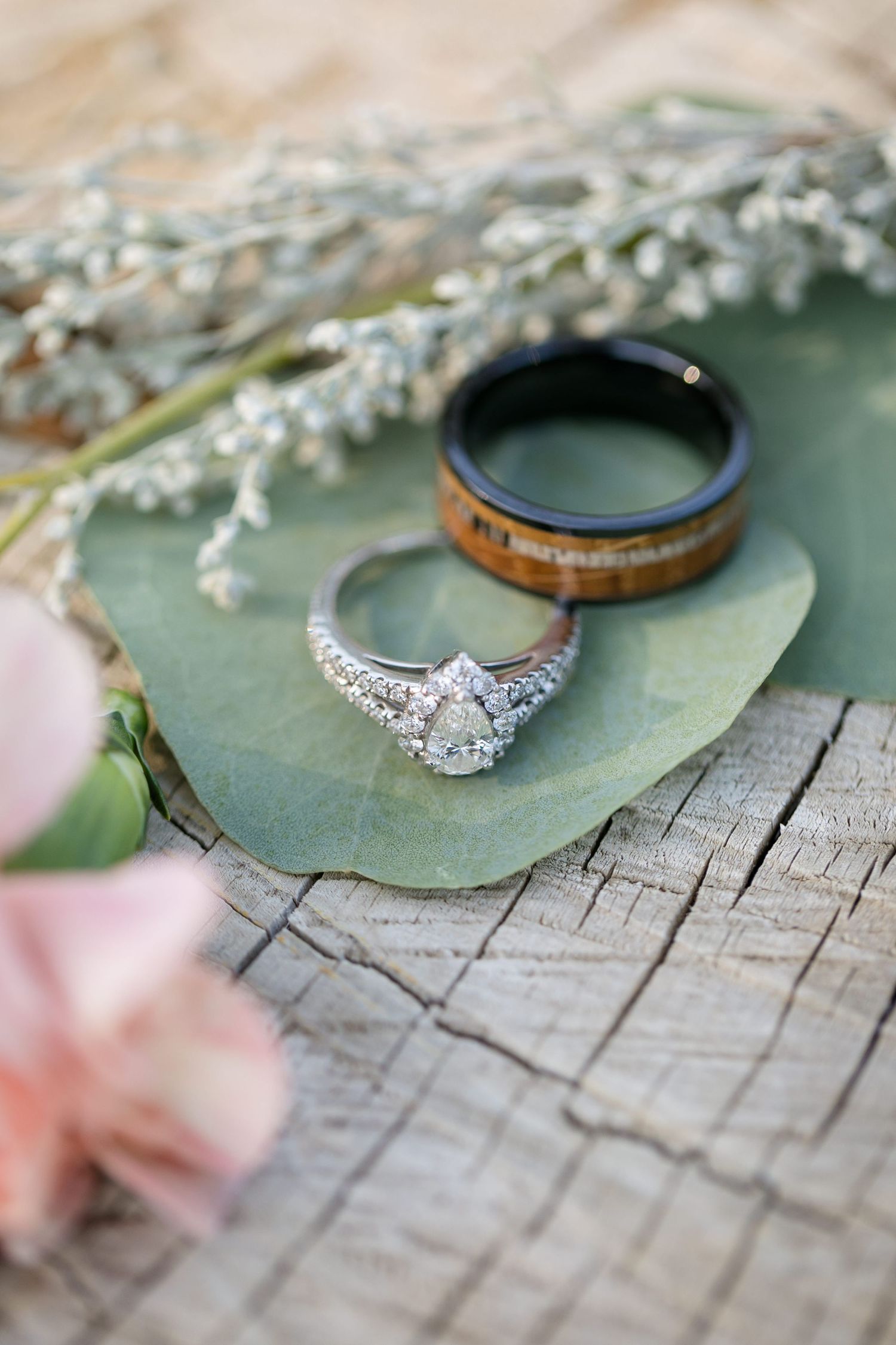 Diamond halo engagement ring and wooden wedding band displayed on eucalyptus leaf with pink flowers and dried botanicals.